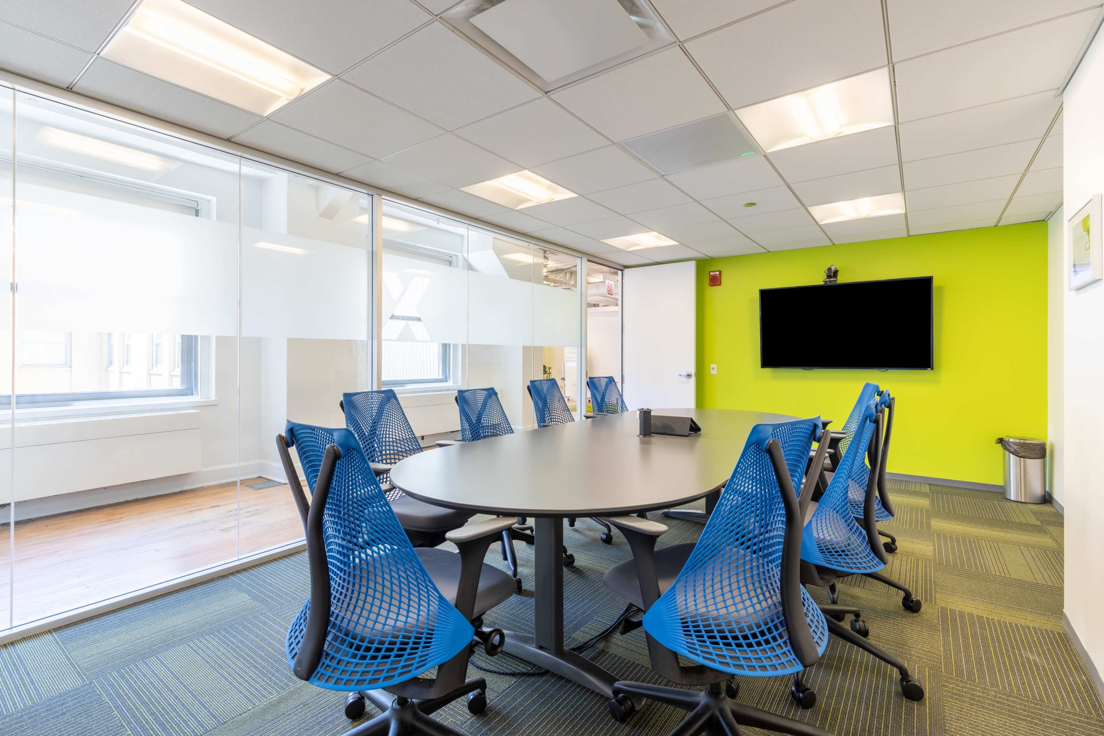 A modern conference room features a round table surrounded by blue mesh chairs, glass walls, and a large television screen mounted on a green accent wall.
