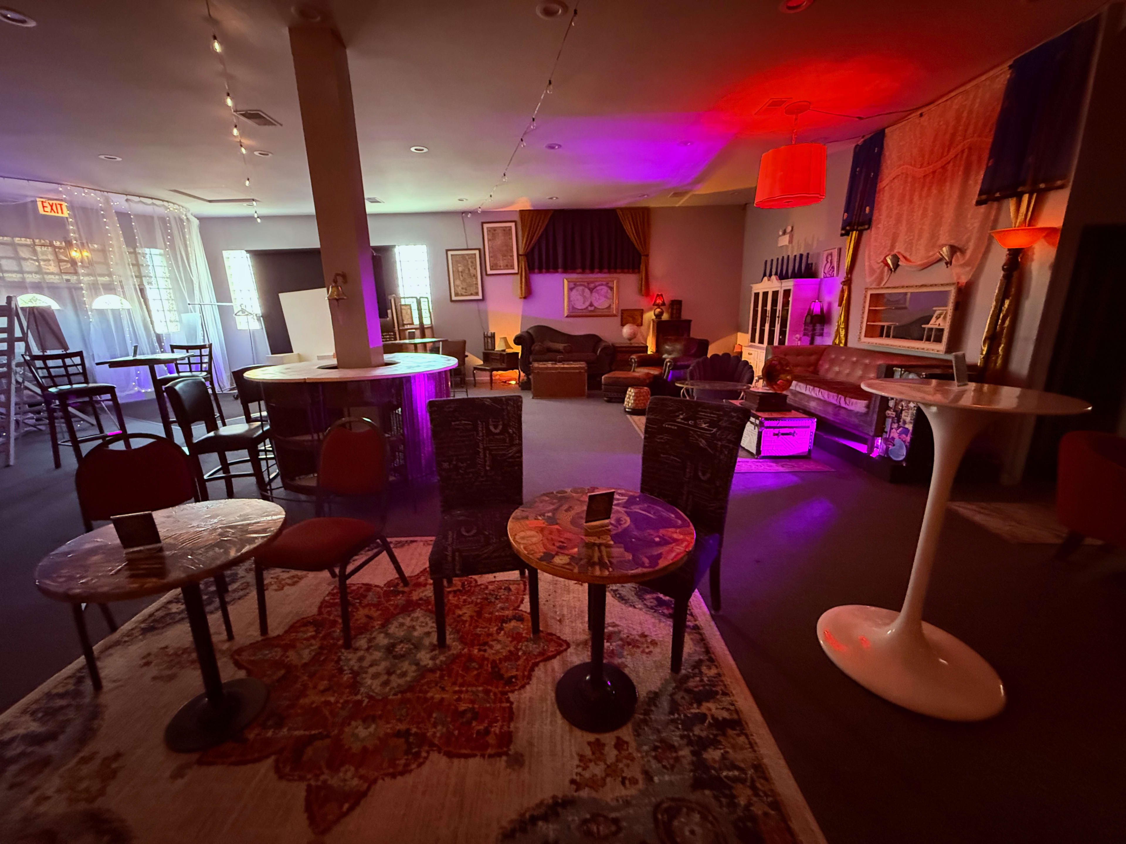 The image shows a dimly lit lounge with various seating arrangements, including tables and chairs, surrounded by decorative elements and soft colored lighting.
