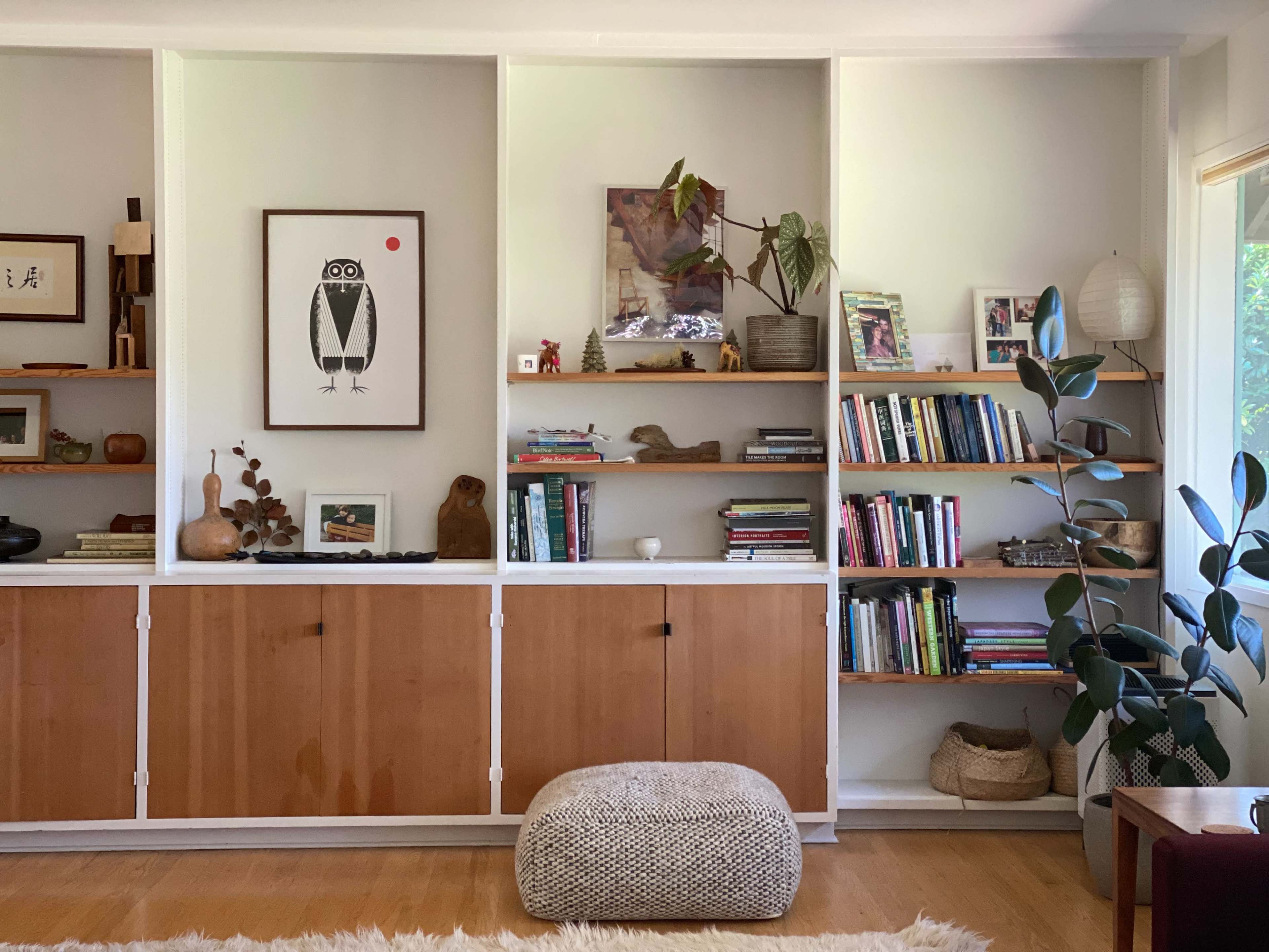 A modern living room with a wooden shelving unit filled with books and decorative items, accompanied by a round pouf and a leafy plant in the foreground.