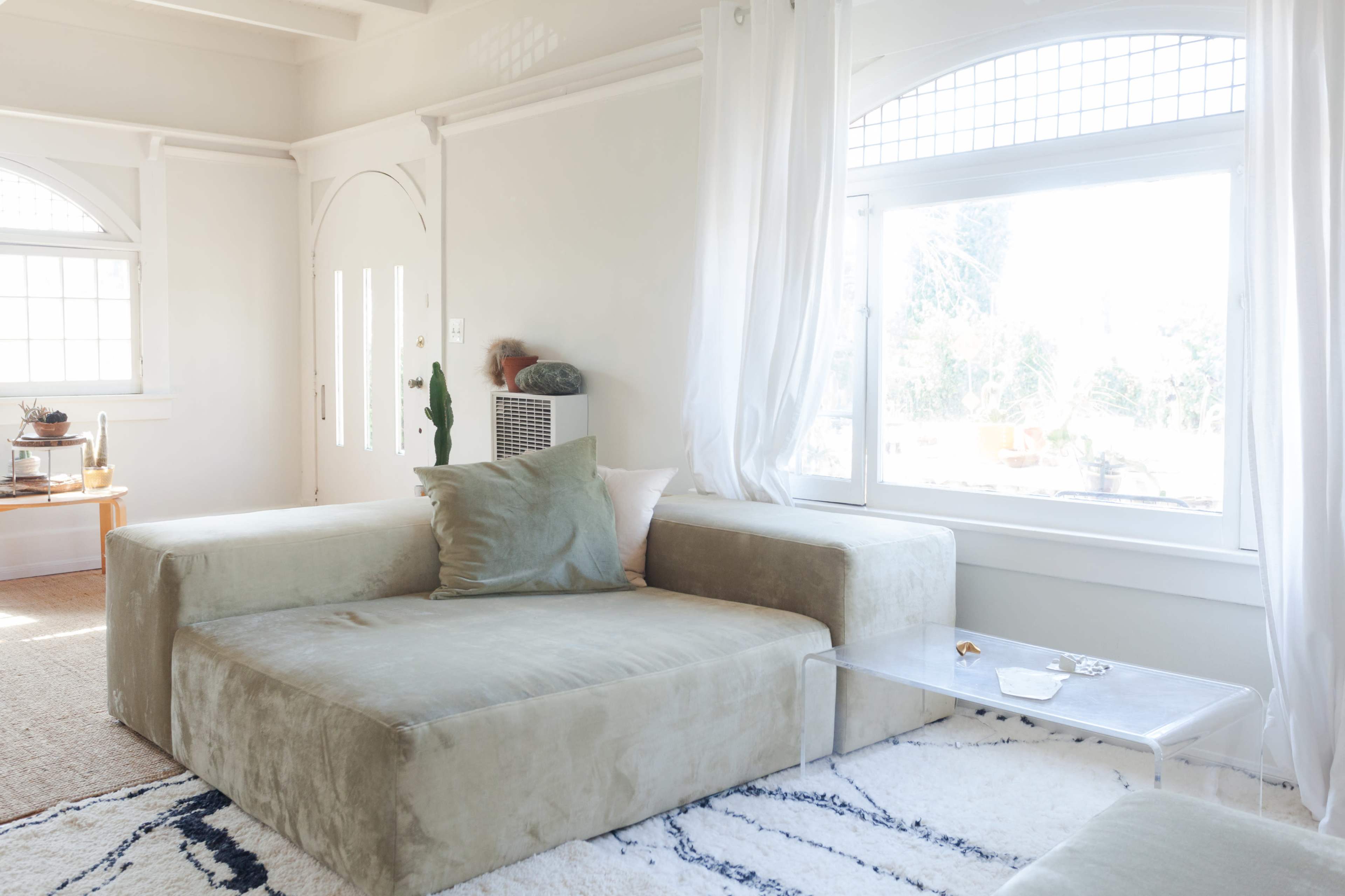The image shows a modern living room with a light green sectional sofa, a clear acrylic side table, and large windows letting in natural light.
