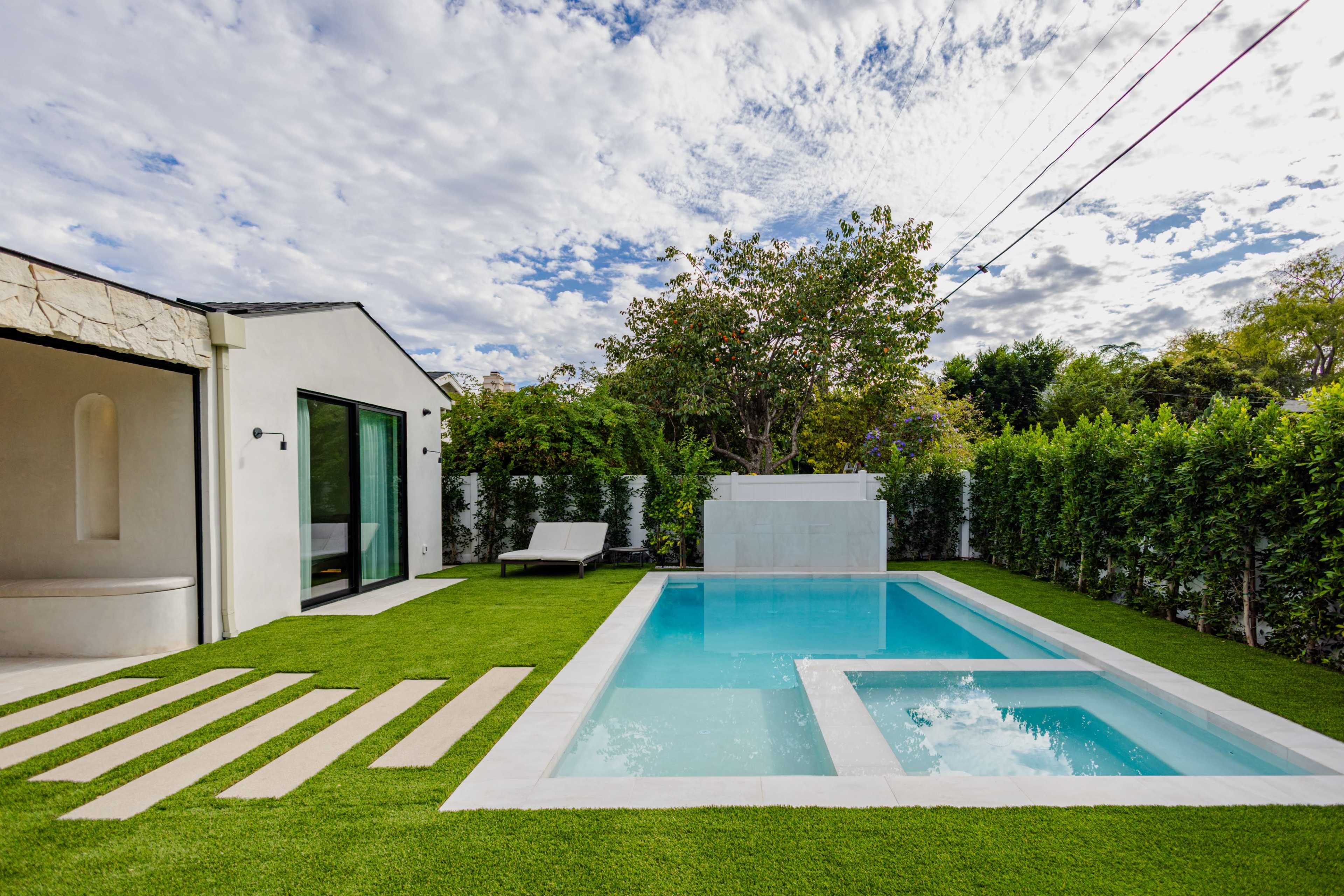 The image shows a modern backyard featuring a rectangular swimming pool surrounded by artificial grass, a concrete pathway, and a small seating area.