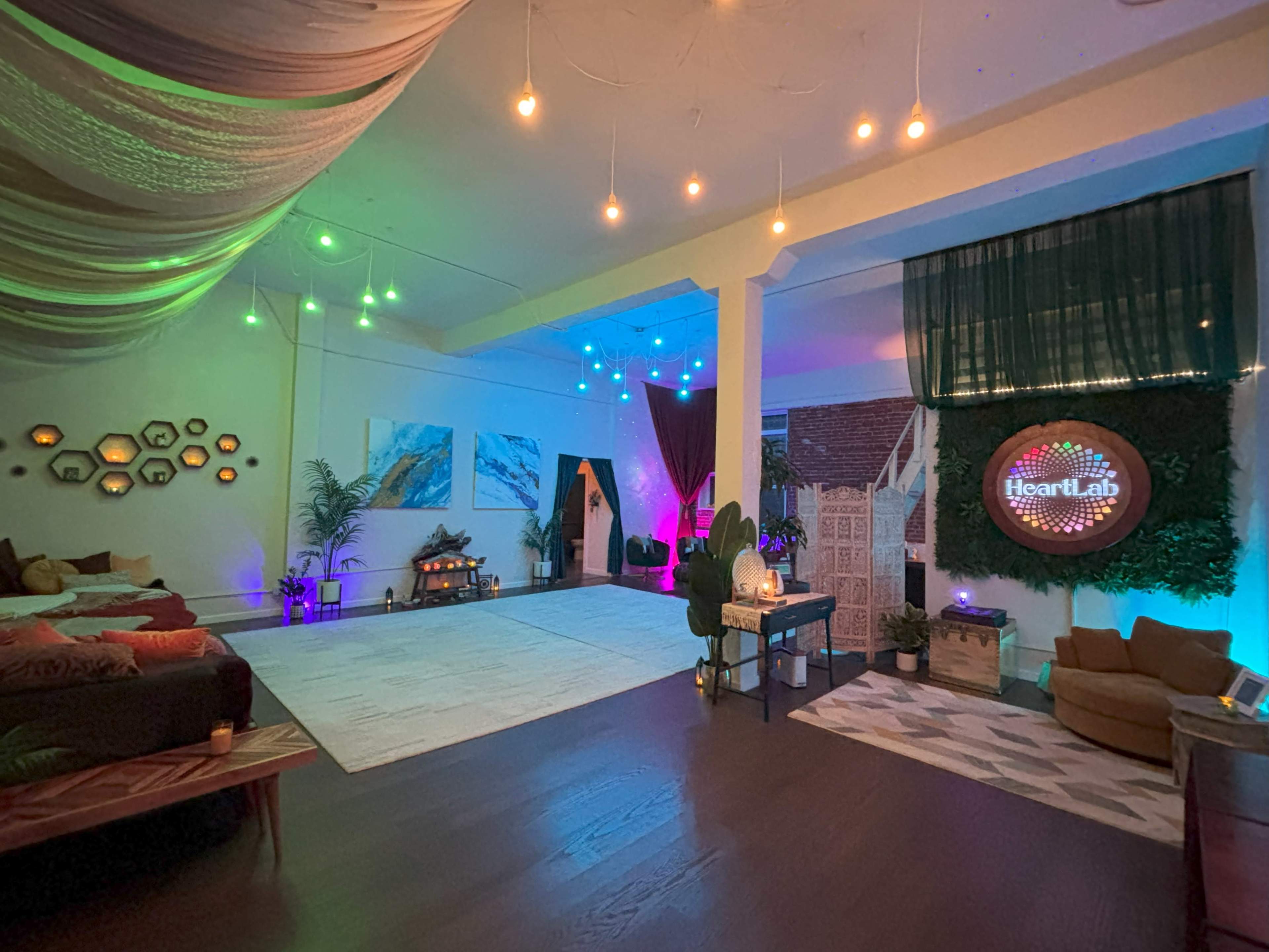 The image shows a cozy, multi-colored room with ambient lighting, featuring comfortable seating, decorative plants, and art on the walls.