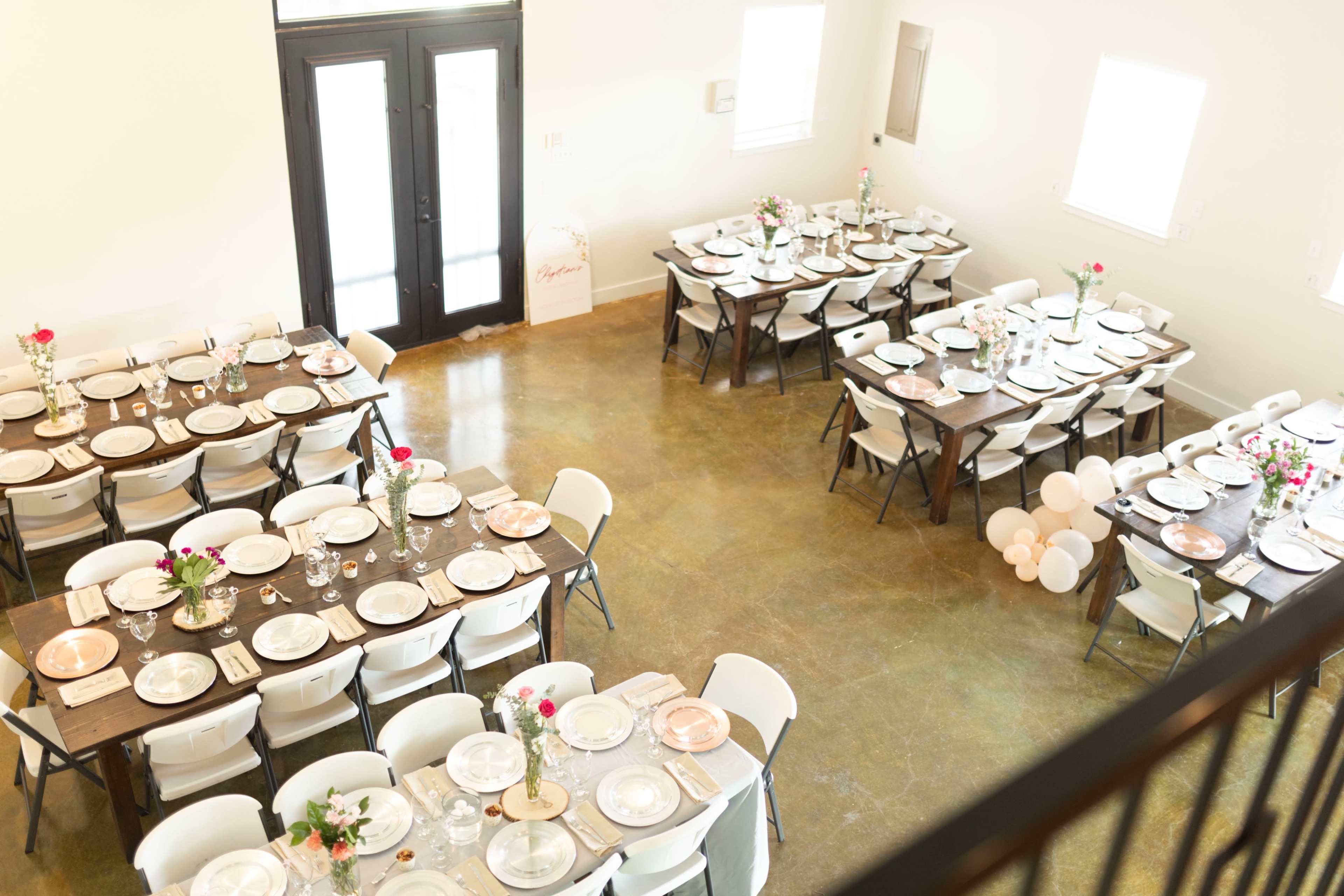 The image shows a spacious dining area arranged with two sets of long tables, each decorated with vases of flowers and white plates, and illuminated by natural light from large windows.