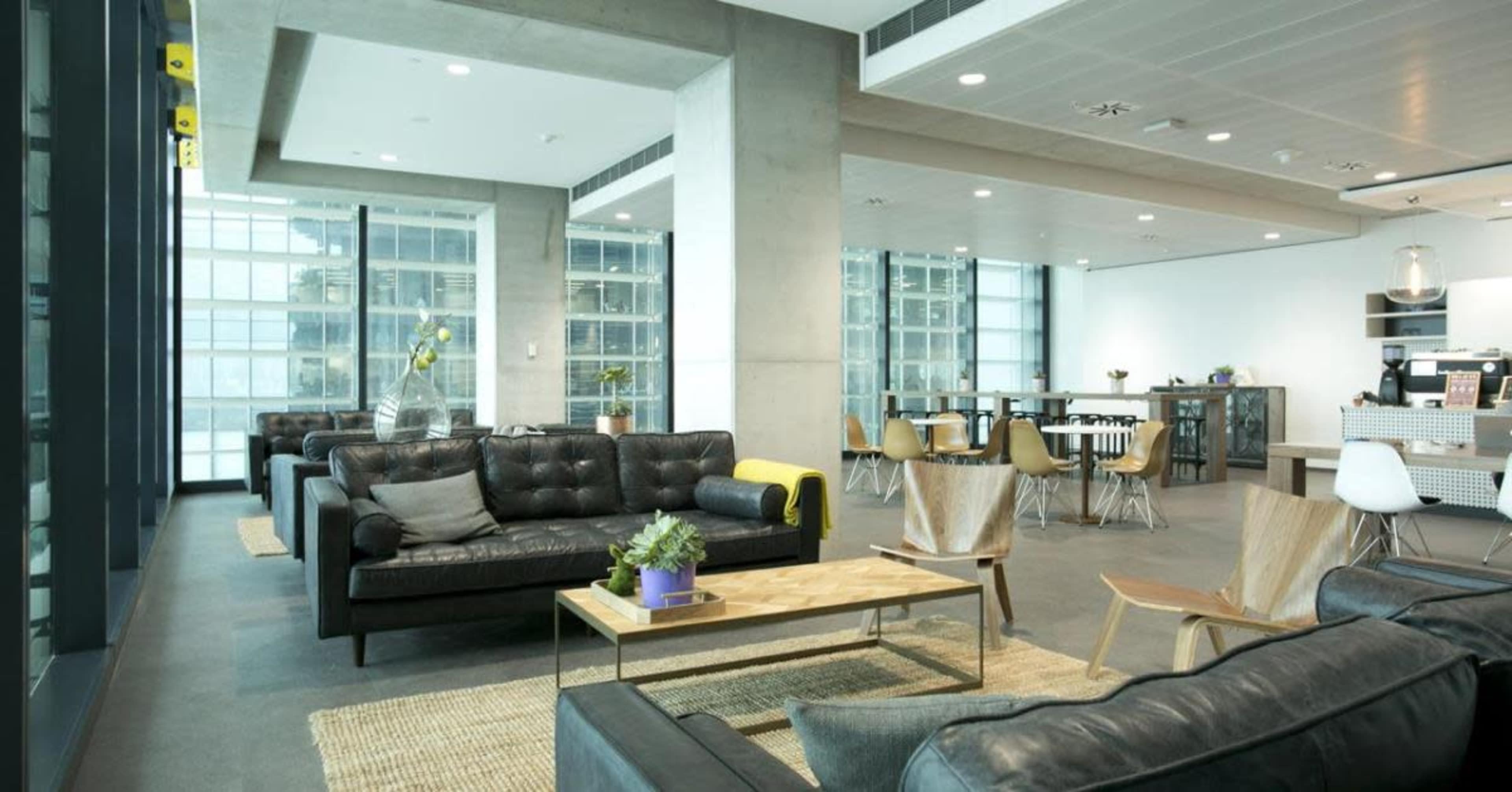 A modern office lounge with dark sofas, a wooden coffee table, and a kitchenette area in the background.