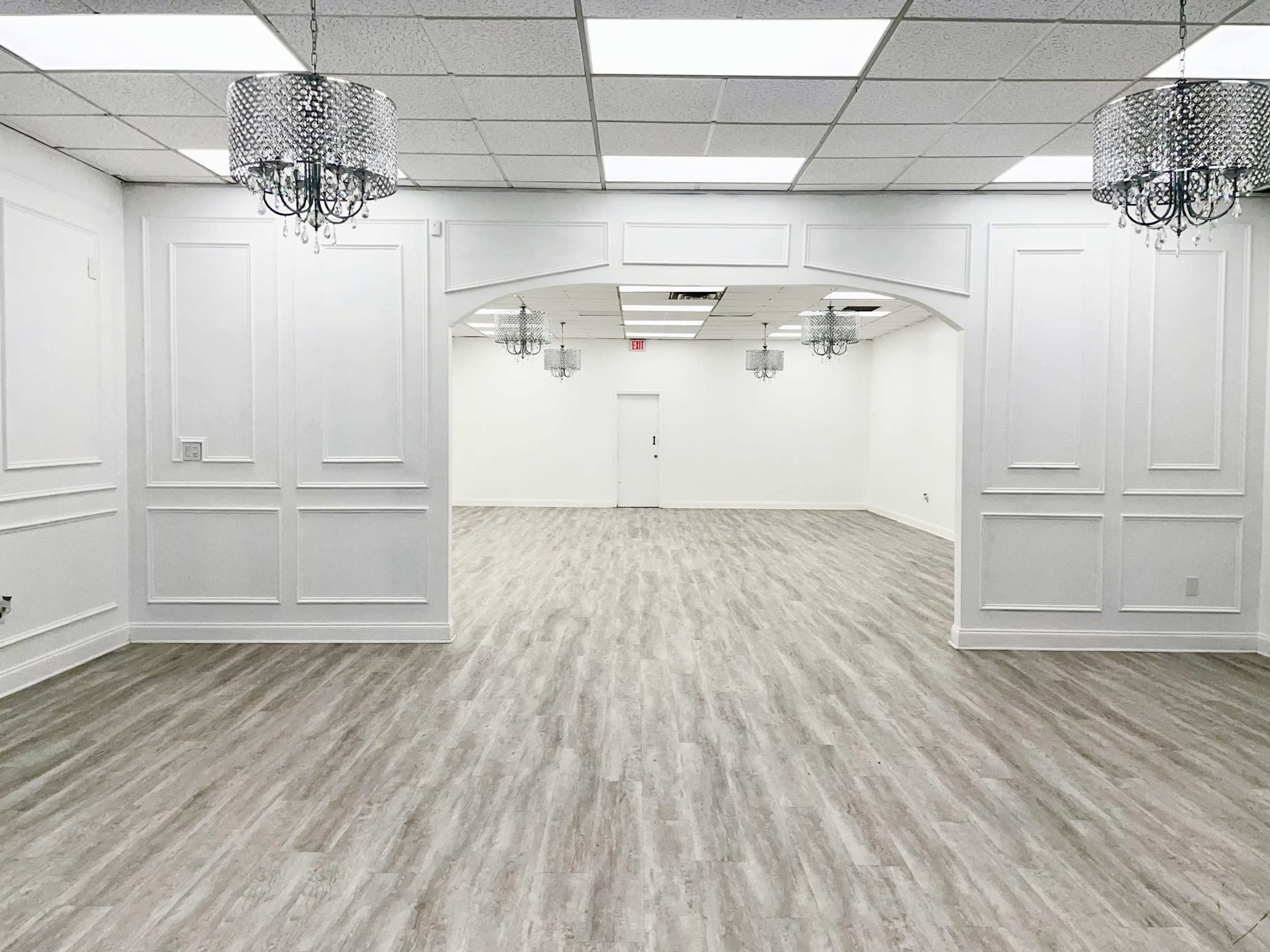 The image shows a large, empty room with white walls and a light wooden floor, featuring several decorative chandeliers hanging from the ceiling.