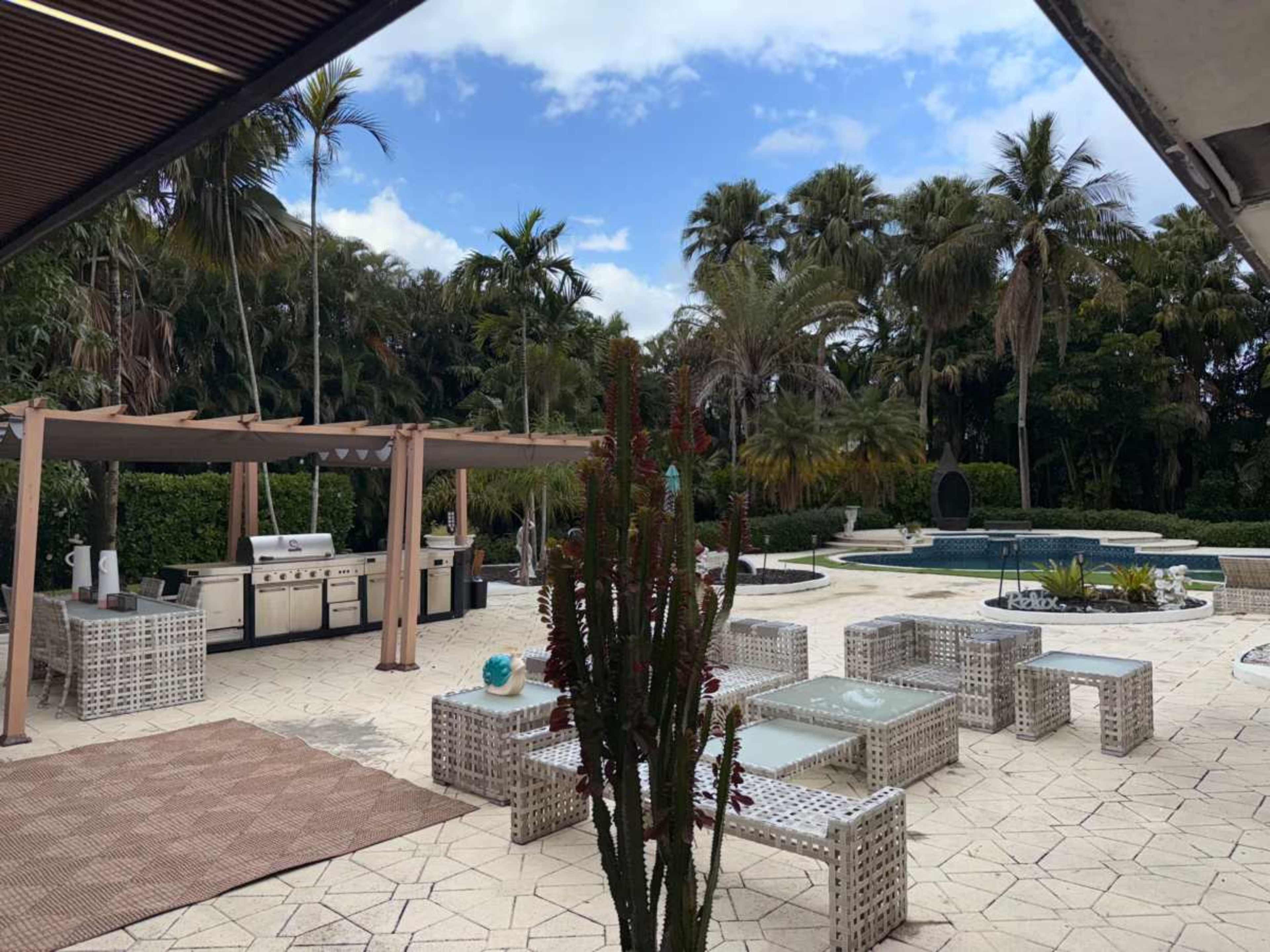 The scene features a spacious outdoor area with a pool, seating made of wicker, and a shaded cooking station surrounded by tropical plants.