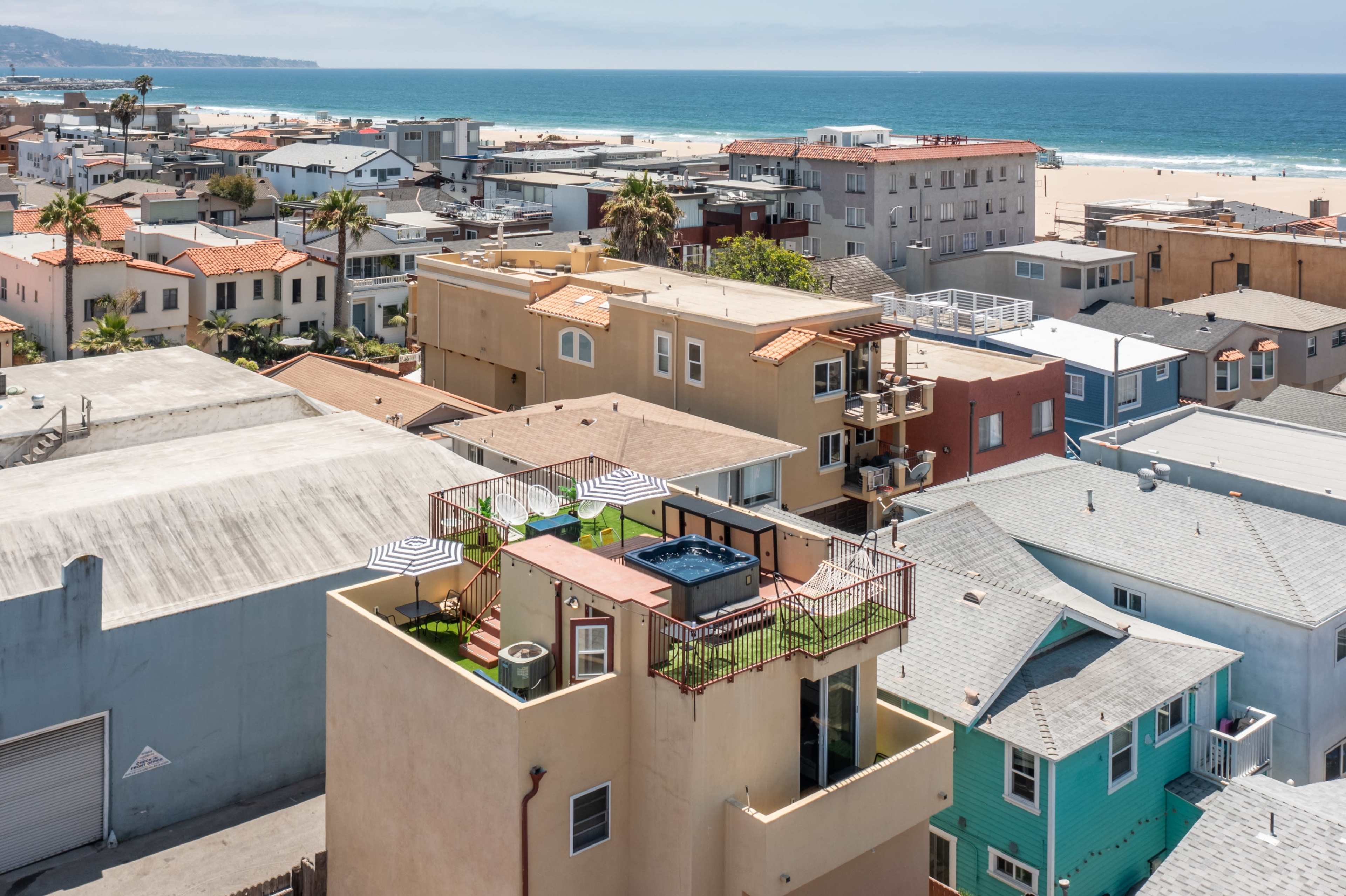 A rooftop deck with a hot tub and lounge chairs is situated atop a multi-story building among a cluster of houses near a beach.