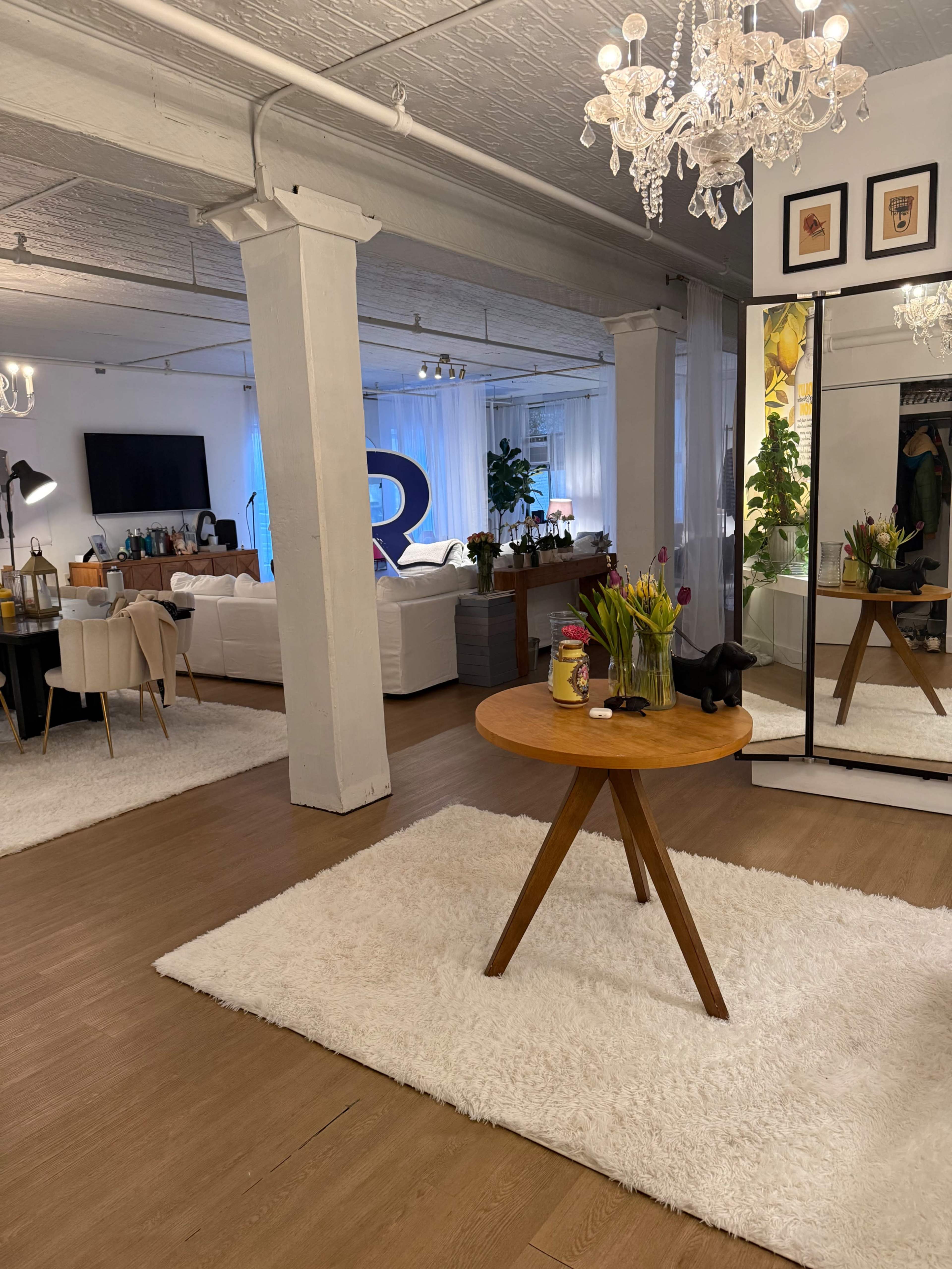 The image shows a stylish interior space with a central round wooden table, a plush white rug, a mirror, and a living area featuring a large couch and a television in the background.
