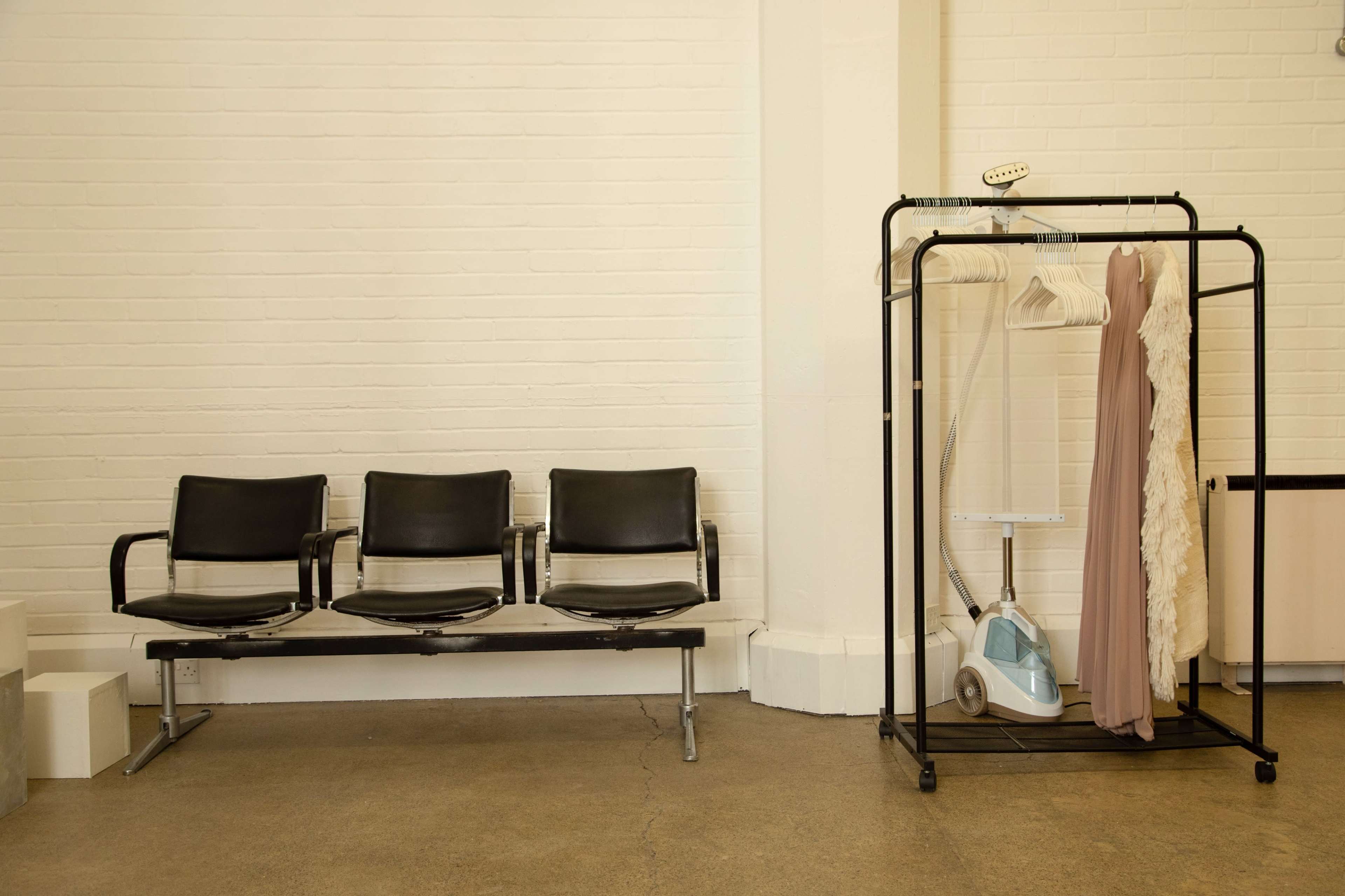 A row of three black seating chairs is positioned next to a clothing rack with a dress and a steamer.