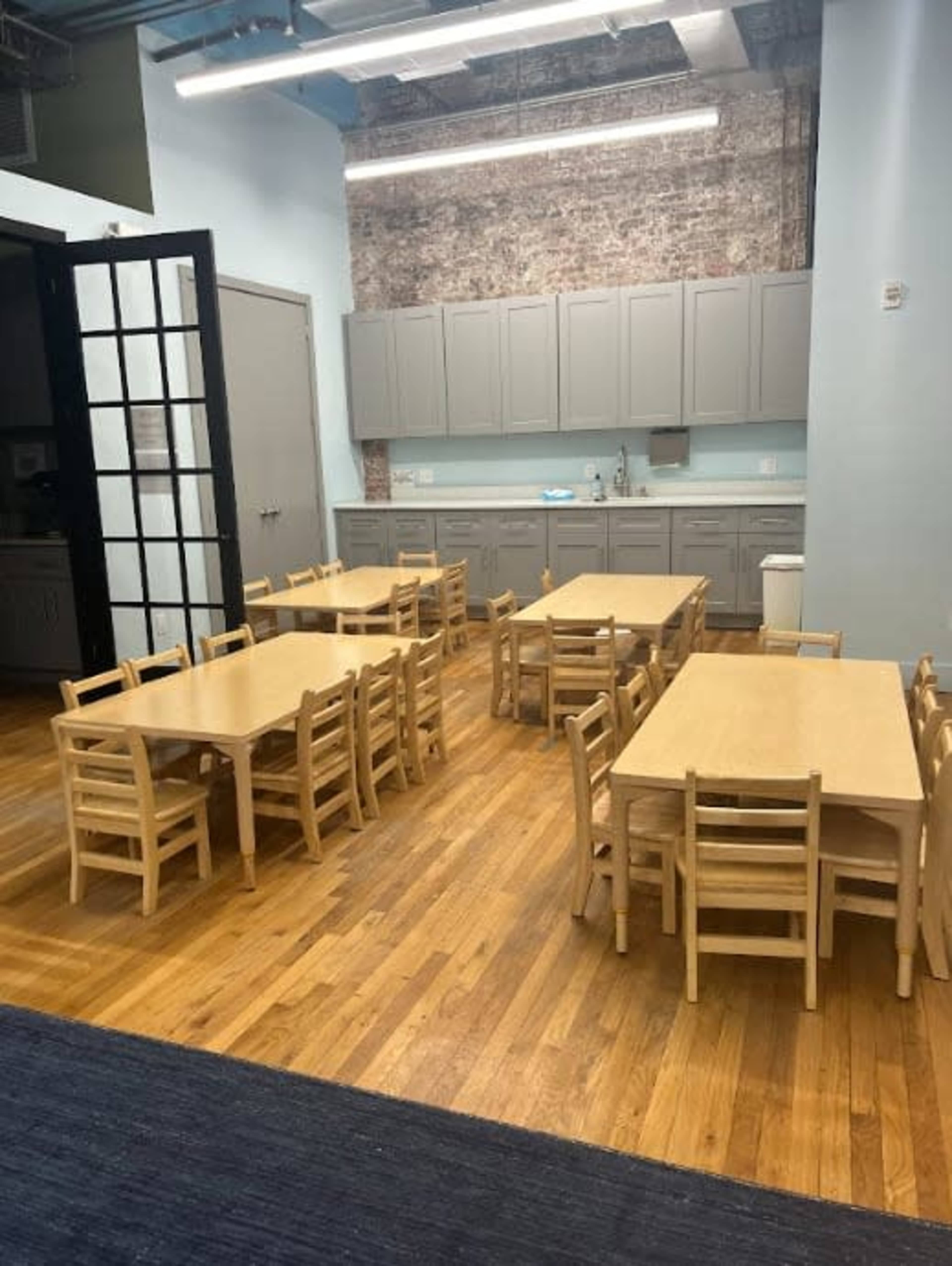 A spacious classroom features multiple wooden tables and chairs arranged neatly, with light blue walls and a kitchenette in the background.