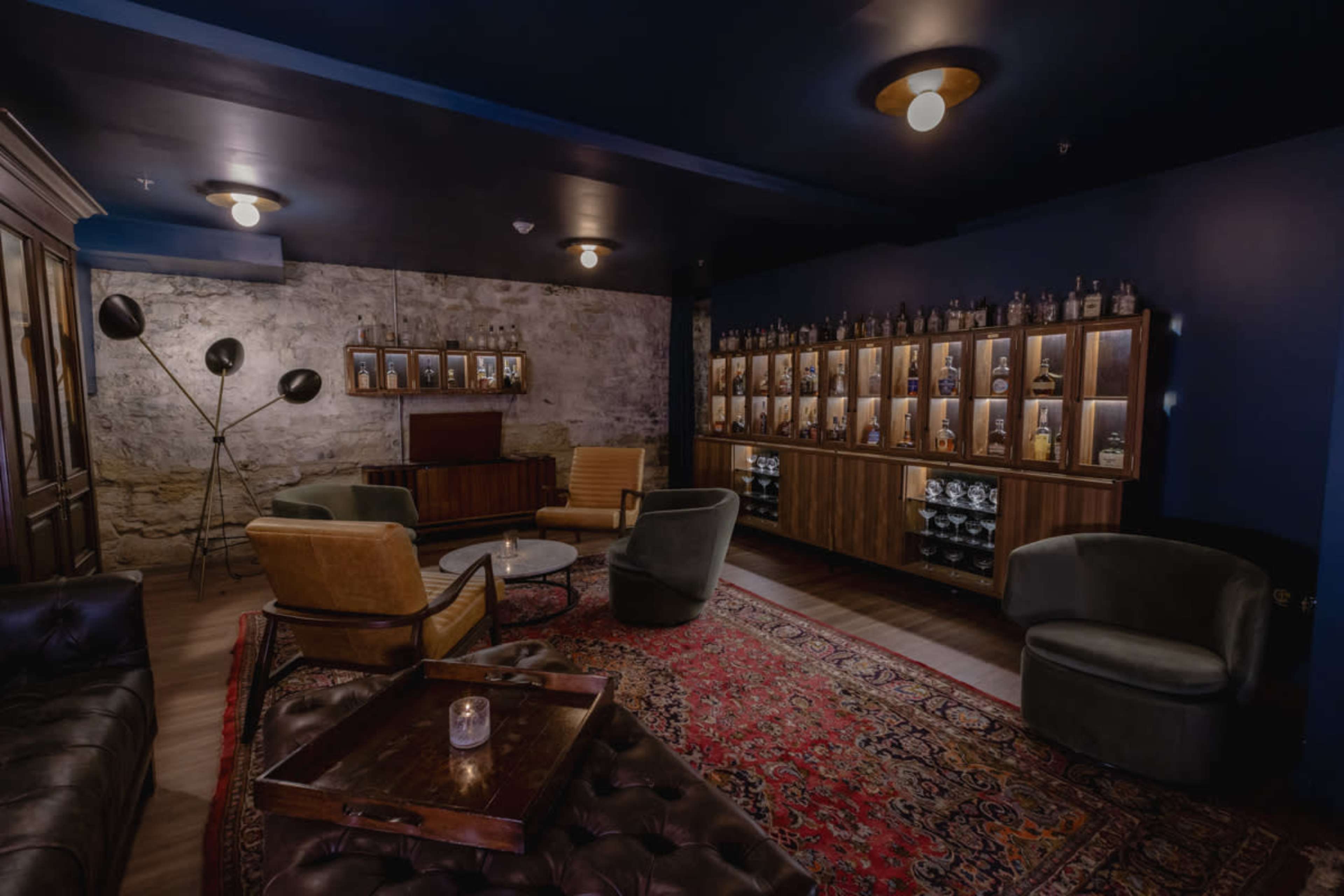 A cozy lounge features dark blue walls, leather seating, a wooden display case filled with bottles, and a large area rug.