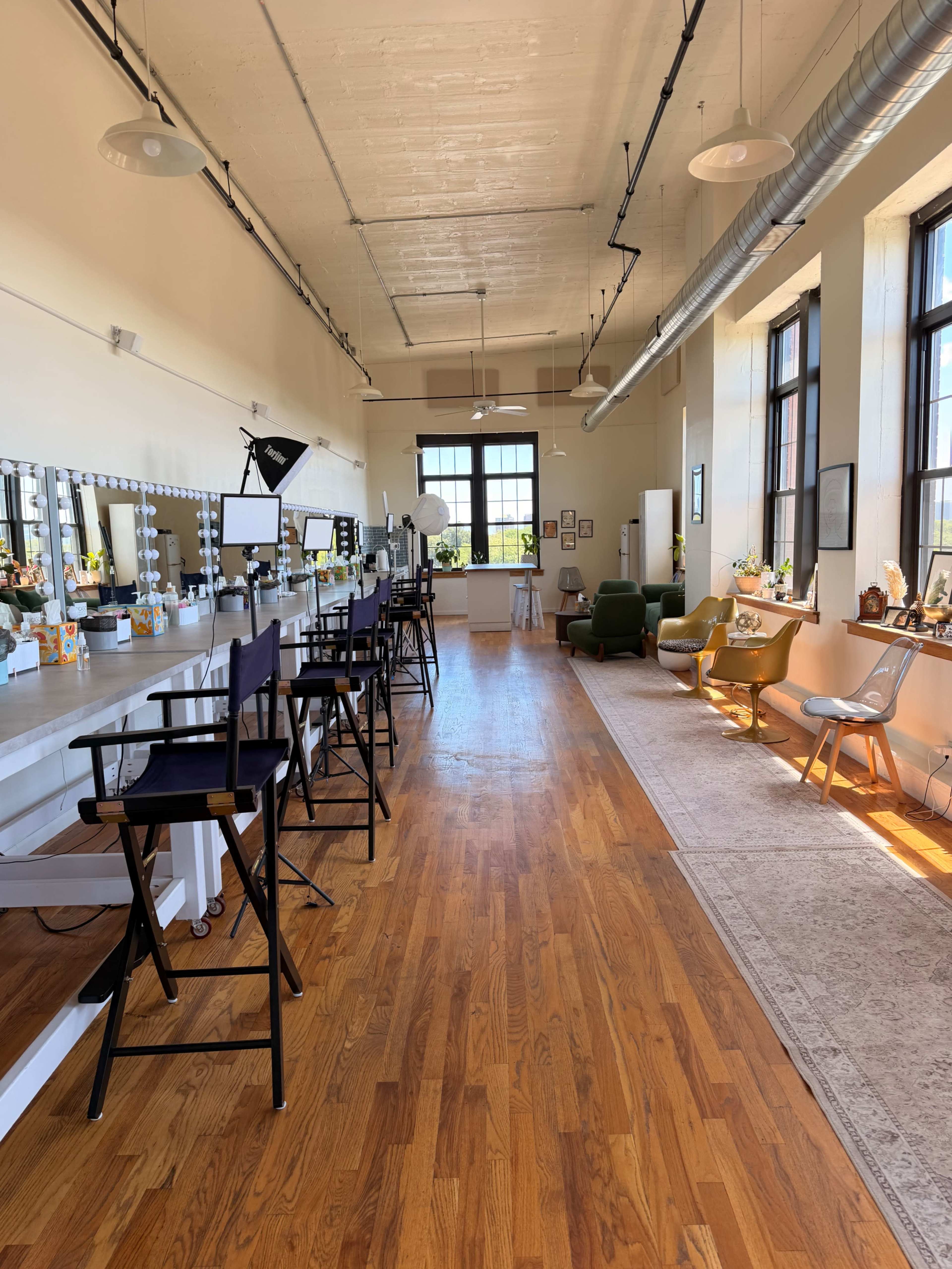 The image shows a spacious, brightly lit studio with makeup chairs along one side and a cozy seating area with various chairs and decor on the opposite side.