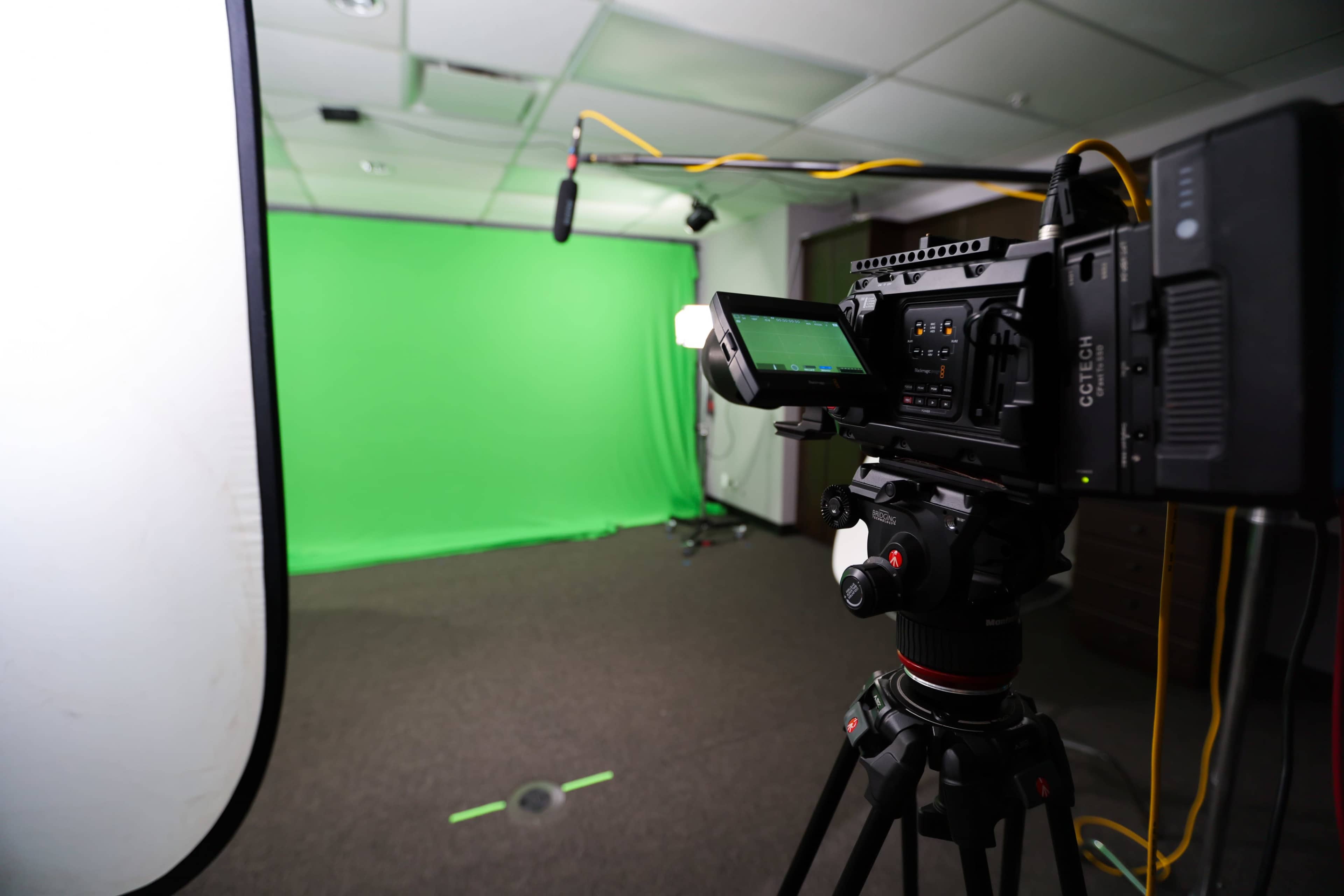 A camera is set up on a tripod facing a green screen in a studio space with lighting and microphones positioned nearby.
