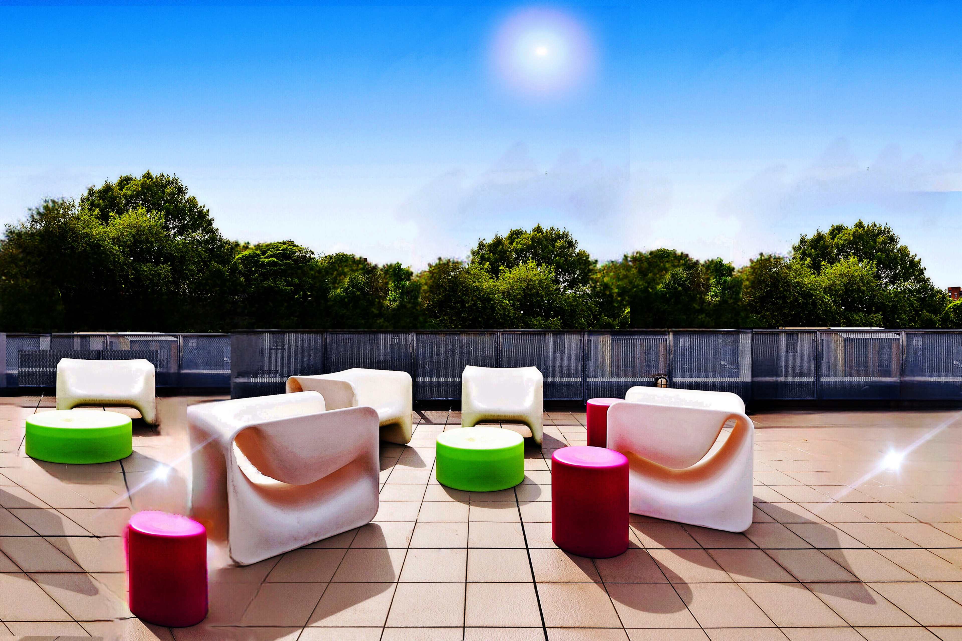 A rooftop area furnished with colorful modern chairs and tables against a clear blue sky and trees in the background.
