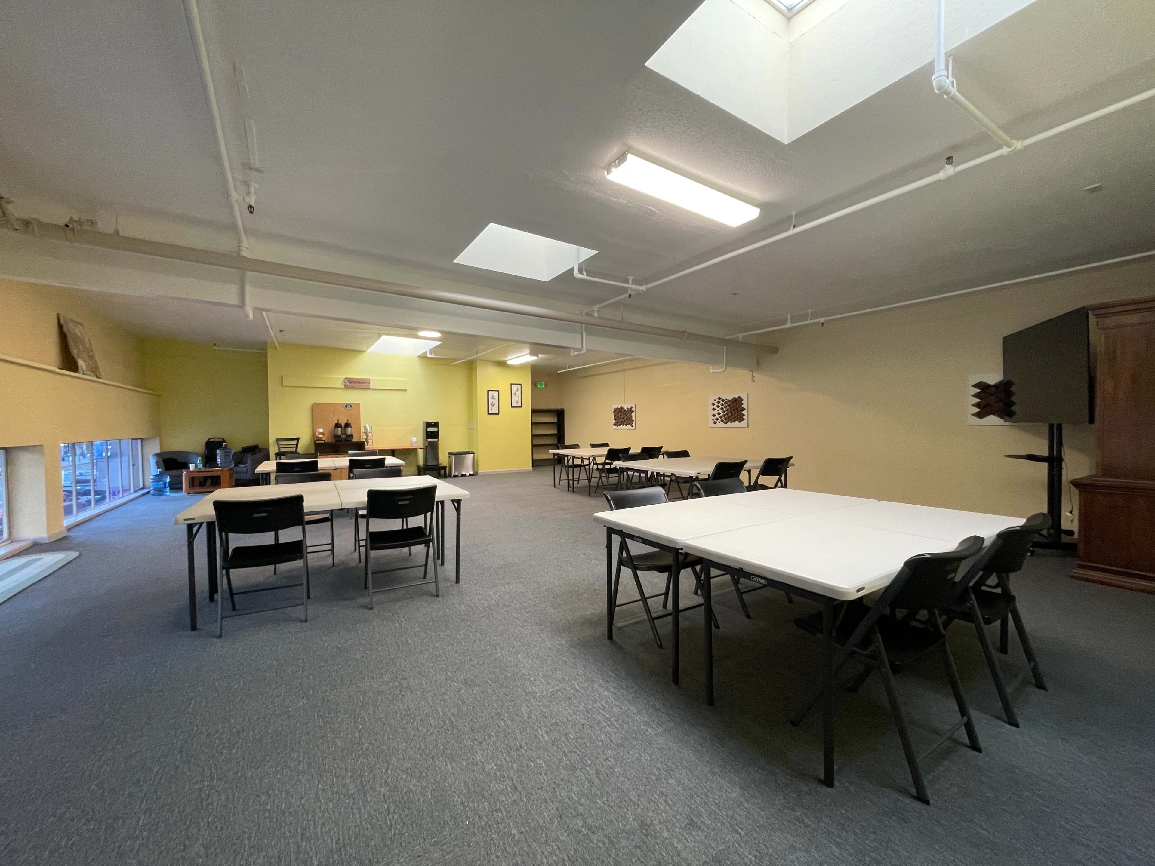 A spacious room with several tables set up, a kitchenette area in the background, and windows allowing natural light to enter.