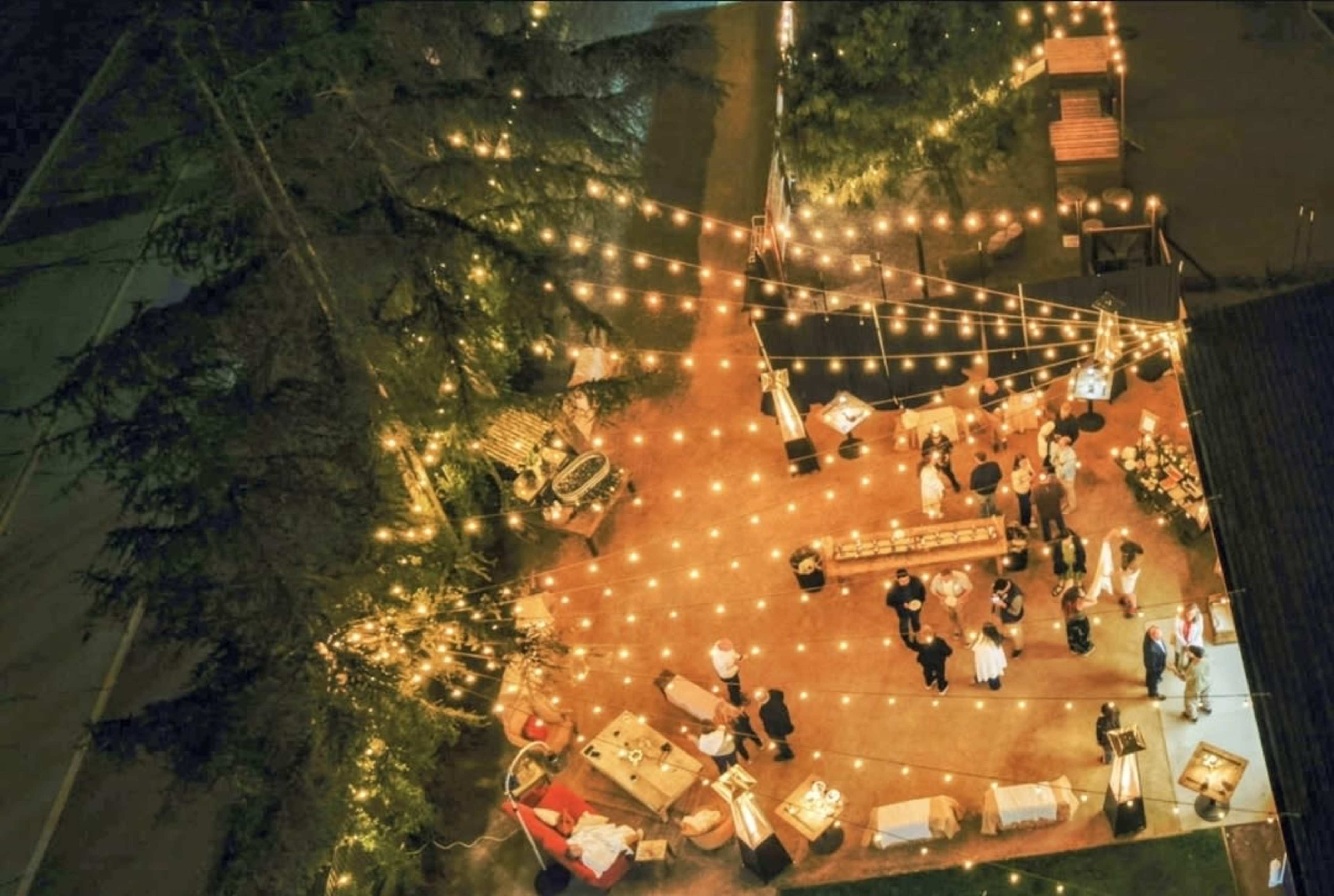 The image shows an outdoor event area illuminated by string lights, with people mingling around food stations and tables.