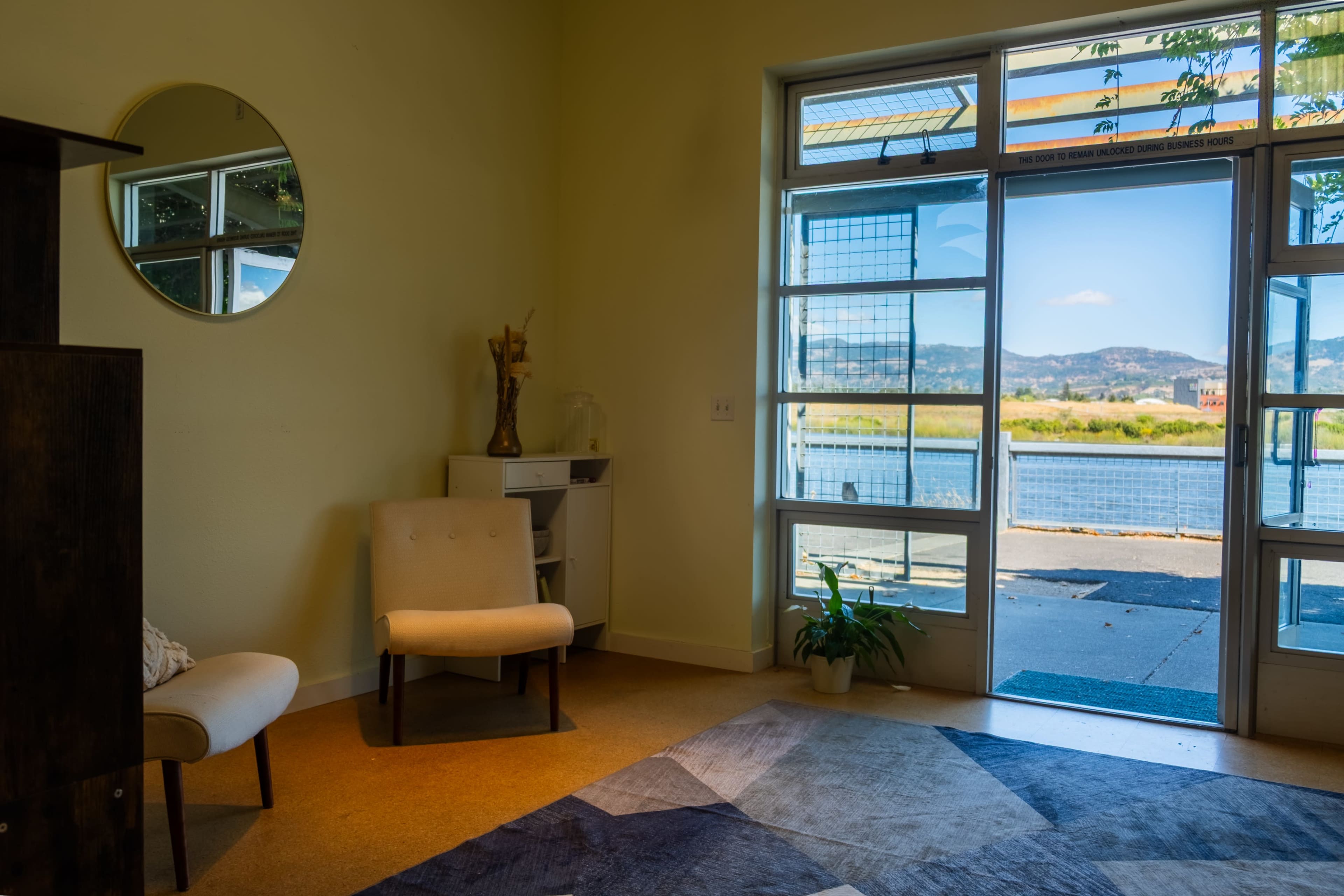 A bright interior space features a chair and small desk, with large windows revealing a view of water and mountains in the background.