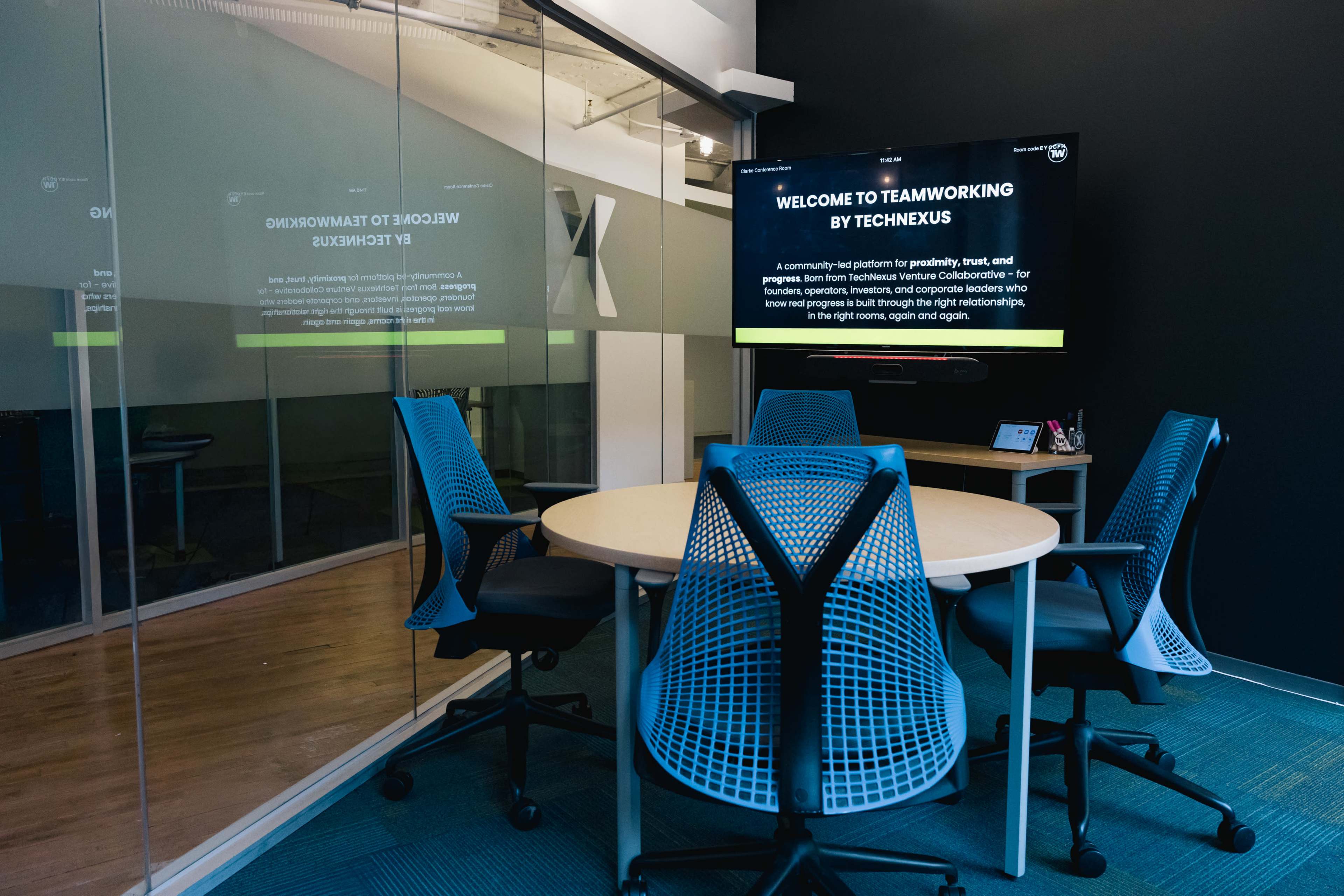 A conference room features a round table surrounded by blue mesh chairs, with a large screen displaying a welcoming message about teamwork.