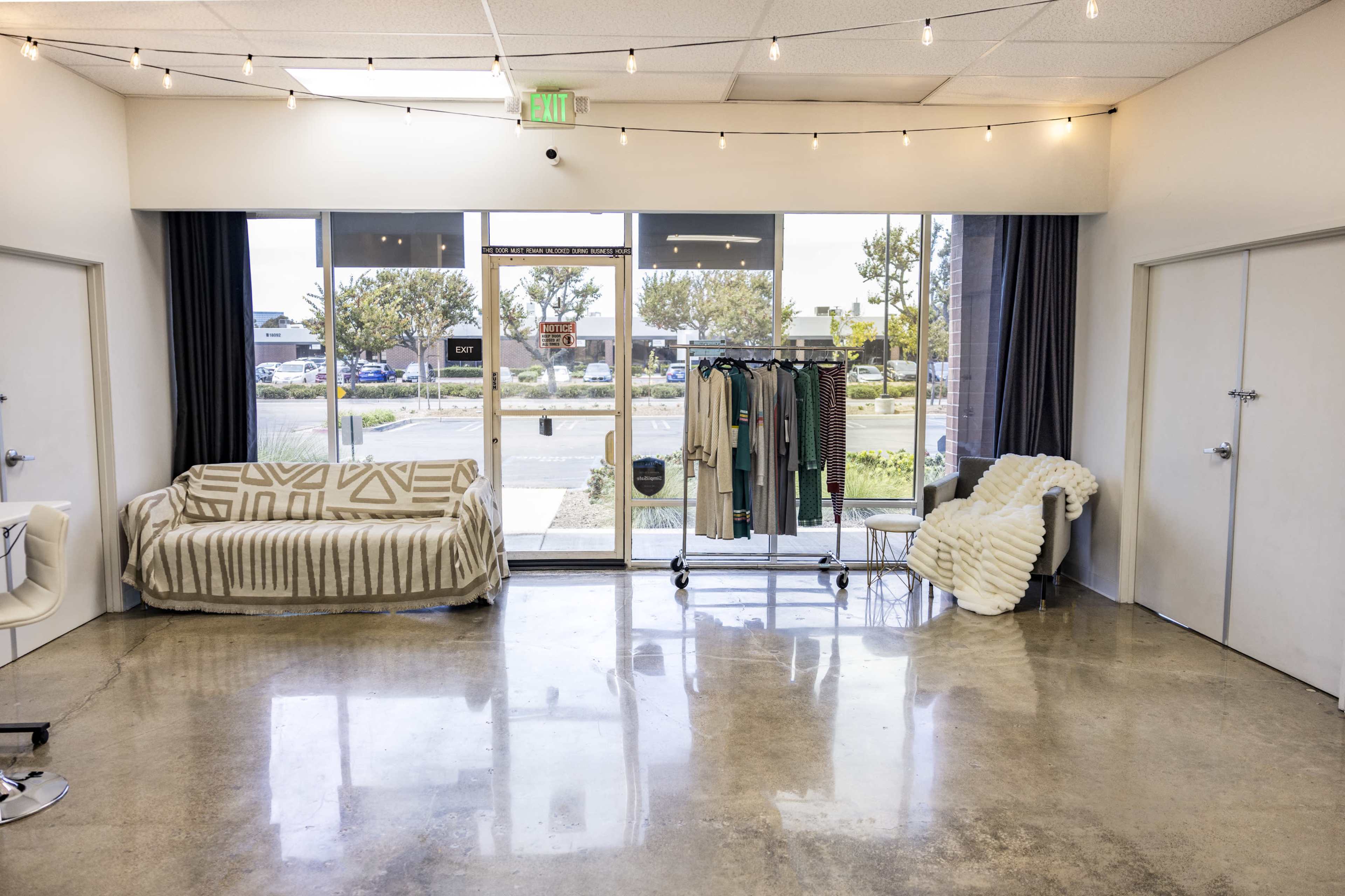 The image shows a vacant retail space with large windows, a sofa, a clothing rack displaying garments, and a cozy chair.