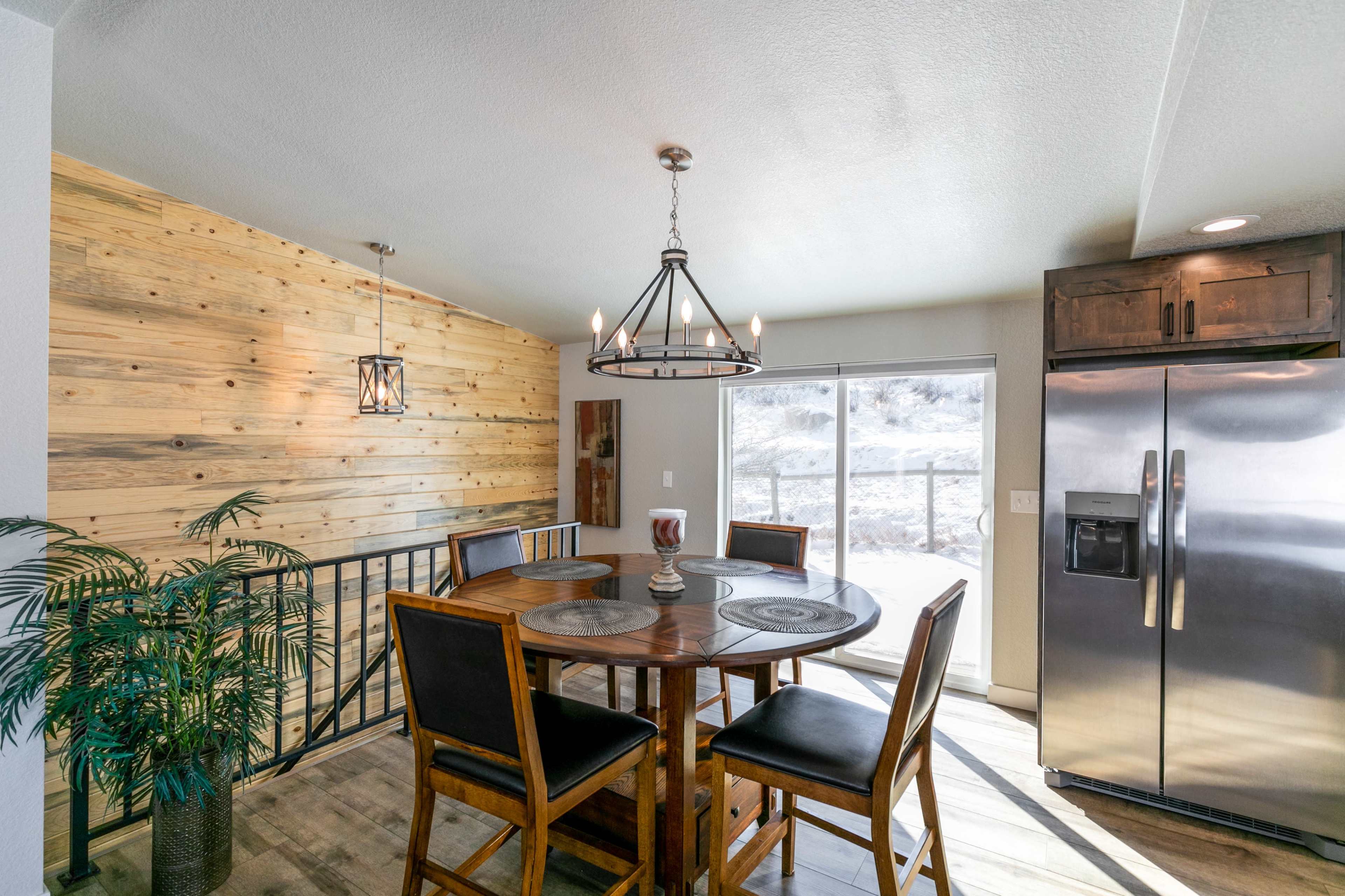 A dining area features a round wooden table with four chairs, set against a wooden accent wall and next to a large window and stainless steel refrigerator.