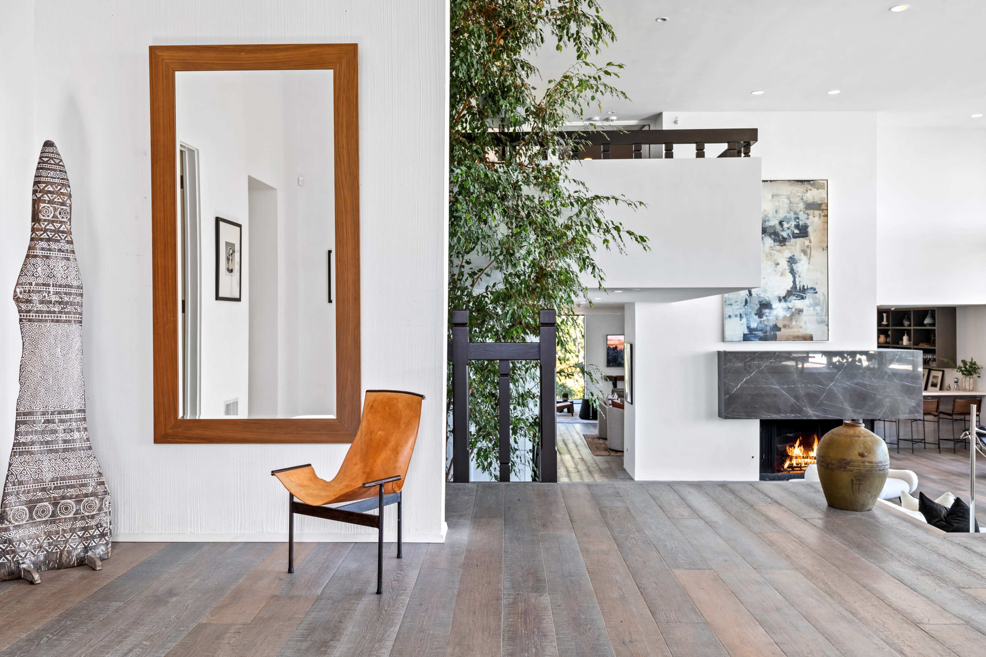 The image shows a spacious indoor area featuring a large wooden-framed mirror, a leather chair, a tall green plant, and a modern fireplace with a stone facade.