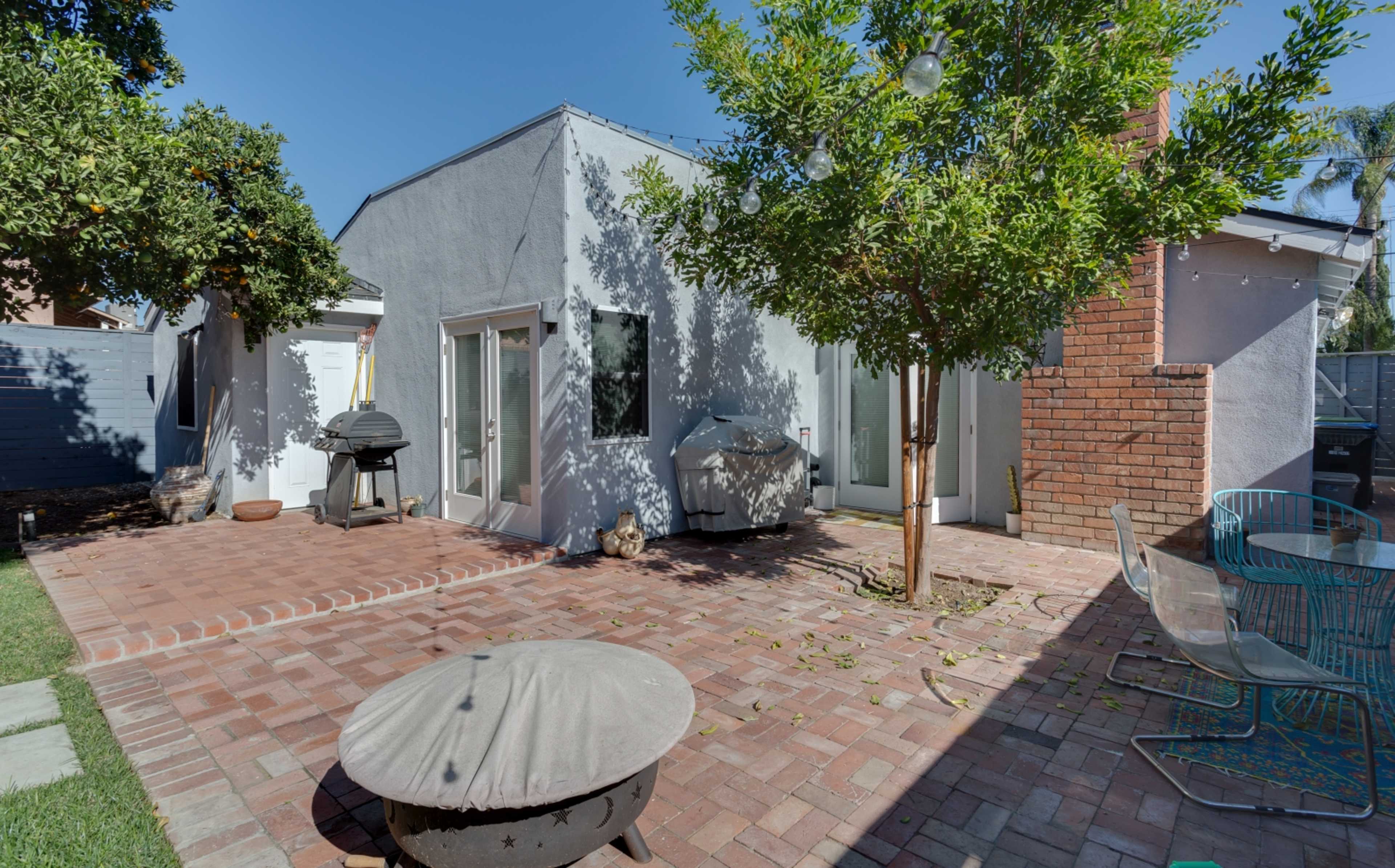 A backyard patio with a brick surface, a grill, and a small tree near a light-colored house.