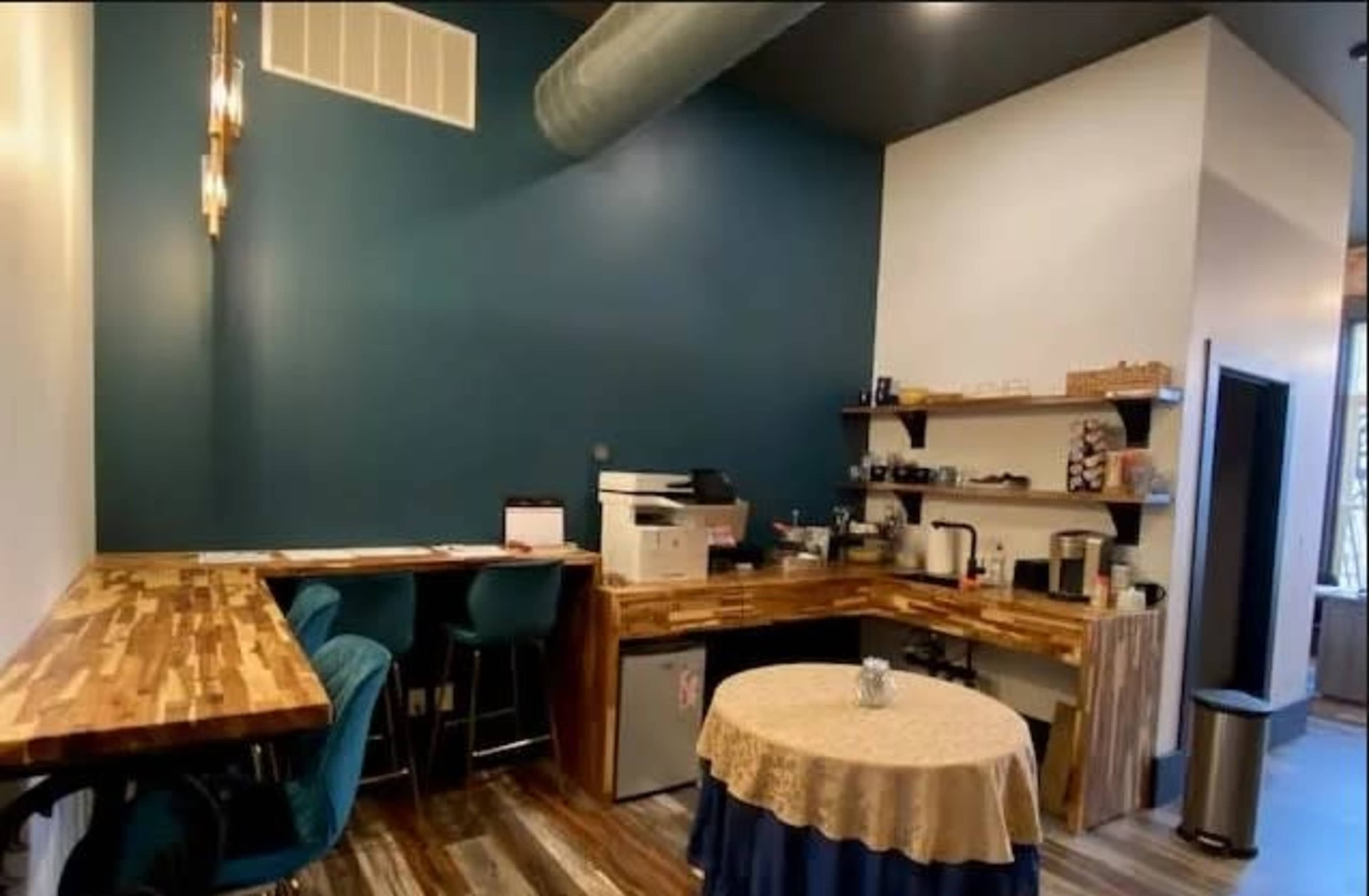 A small coffee shop interior features a wooden counter, teal walls, a shelf with coffee equipment, and a table covered with a tablecloth.