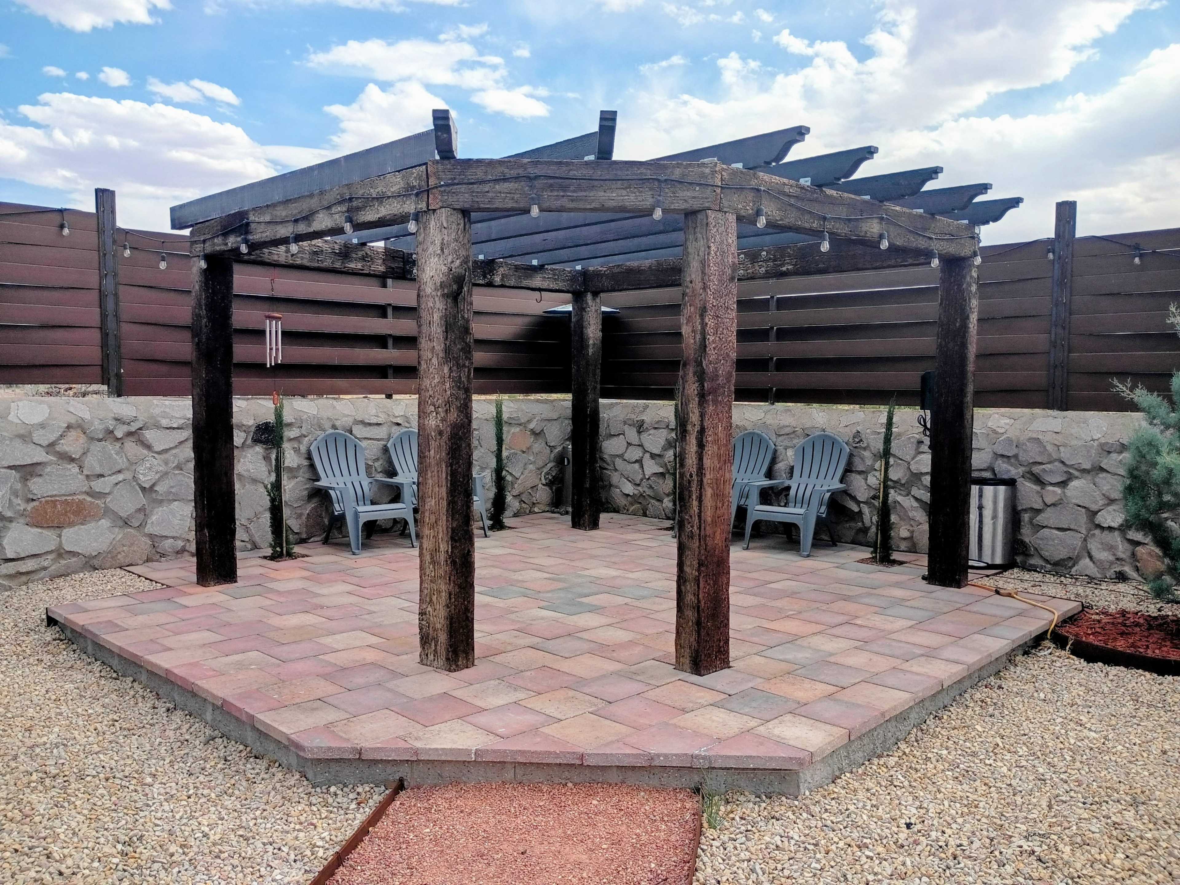 The image shows a patio area with a wooden pergola supported by four posts, surrounded by stone walls and featuring two blue chairs on a patterned stone floor.