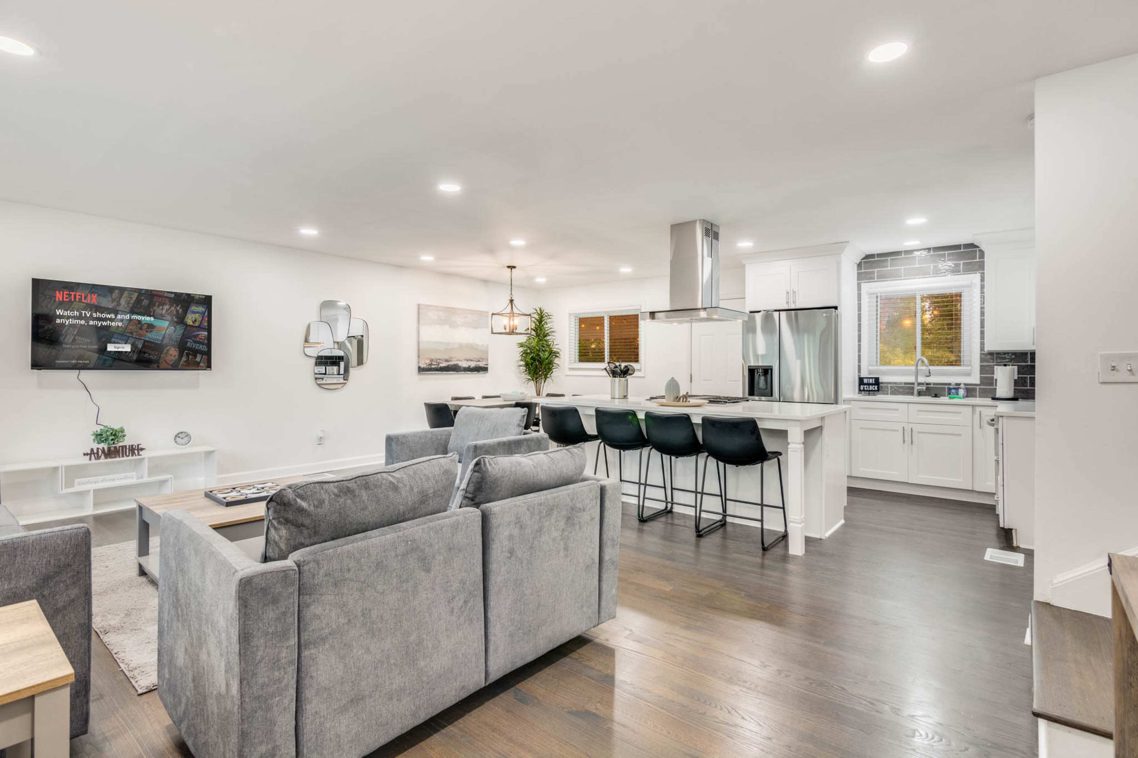 The image shows a modern living room and kitchen area, featuring gray sofas, a dining bar with black stools, and a television mounted on the wall.