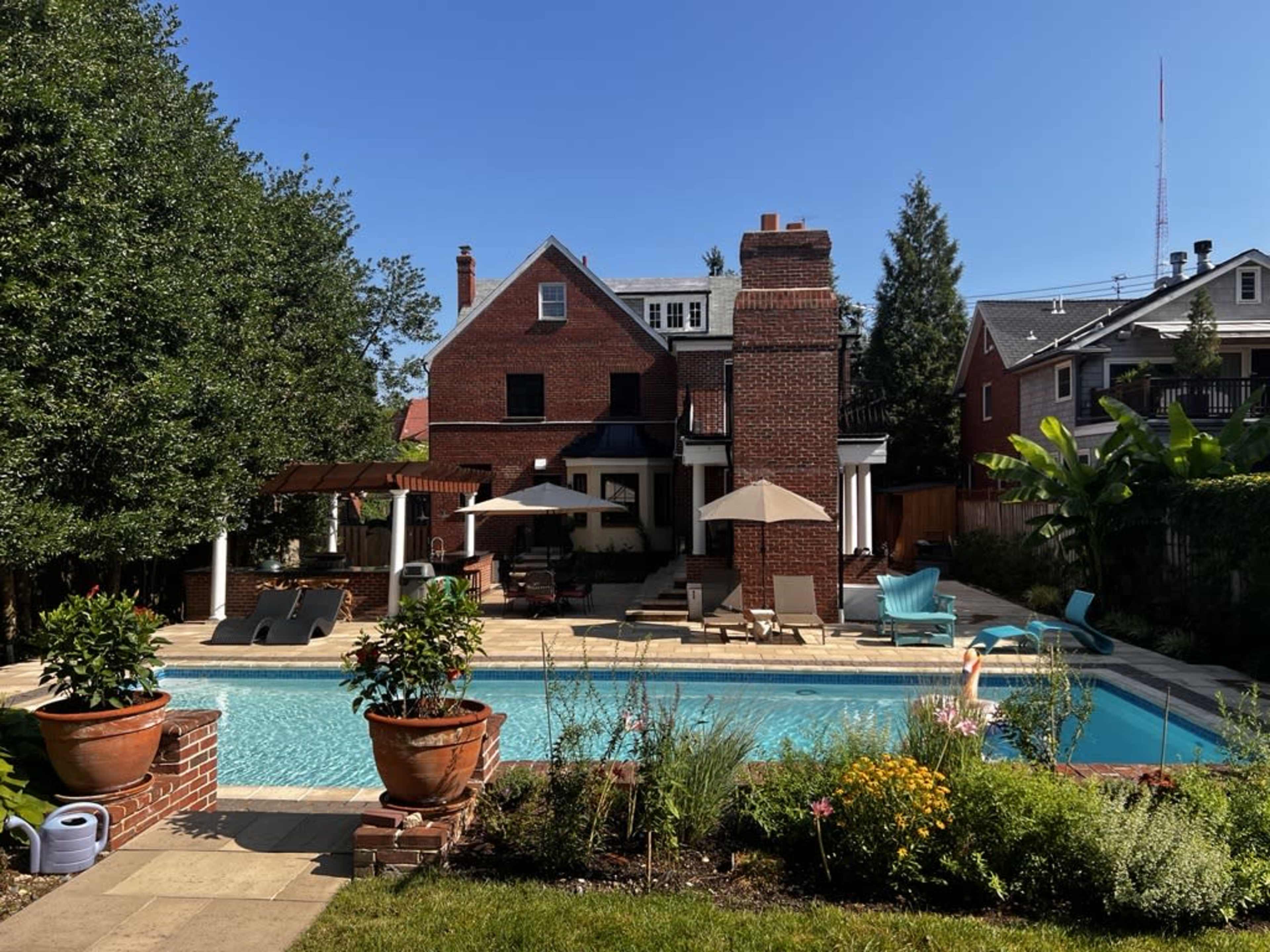 A backyard pool area surrounded by a brick house, lounge chairs, umbrellas, and landscaped greenery.
