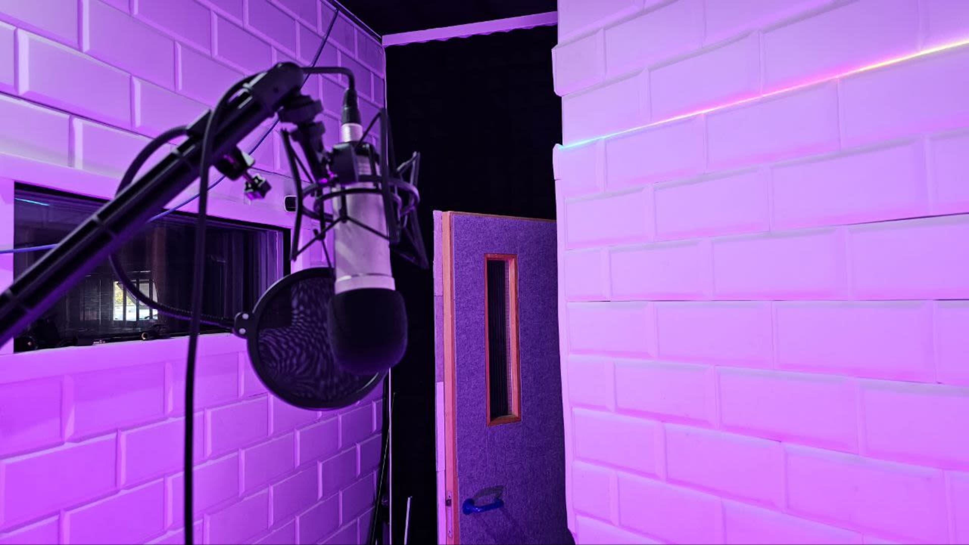 A recording studio with a microphone on a stand and an open door leading to another room, illuminated by purple lighting.