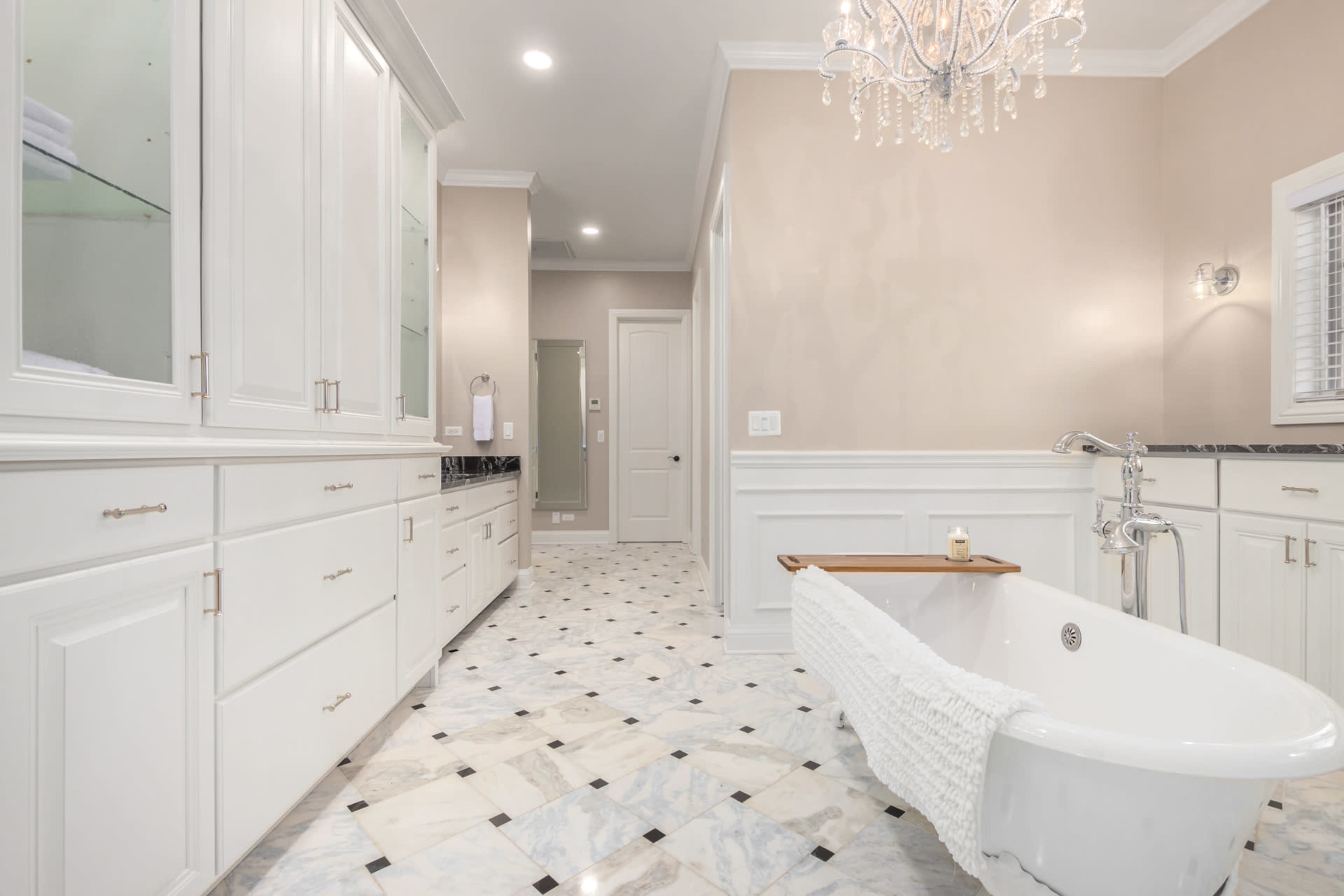 A spacious bathroom with a freestanding bathtub, elegant cabinetry, marble flooring, and a chandelier.