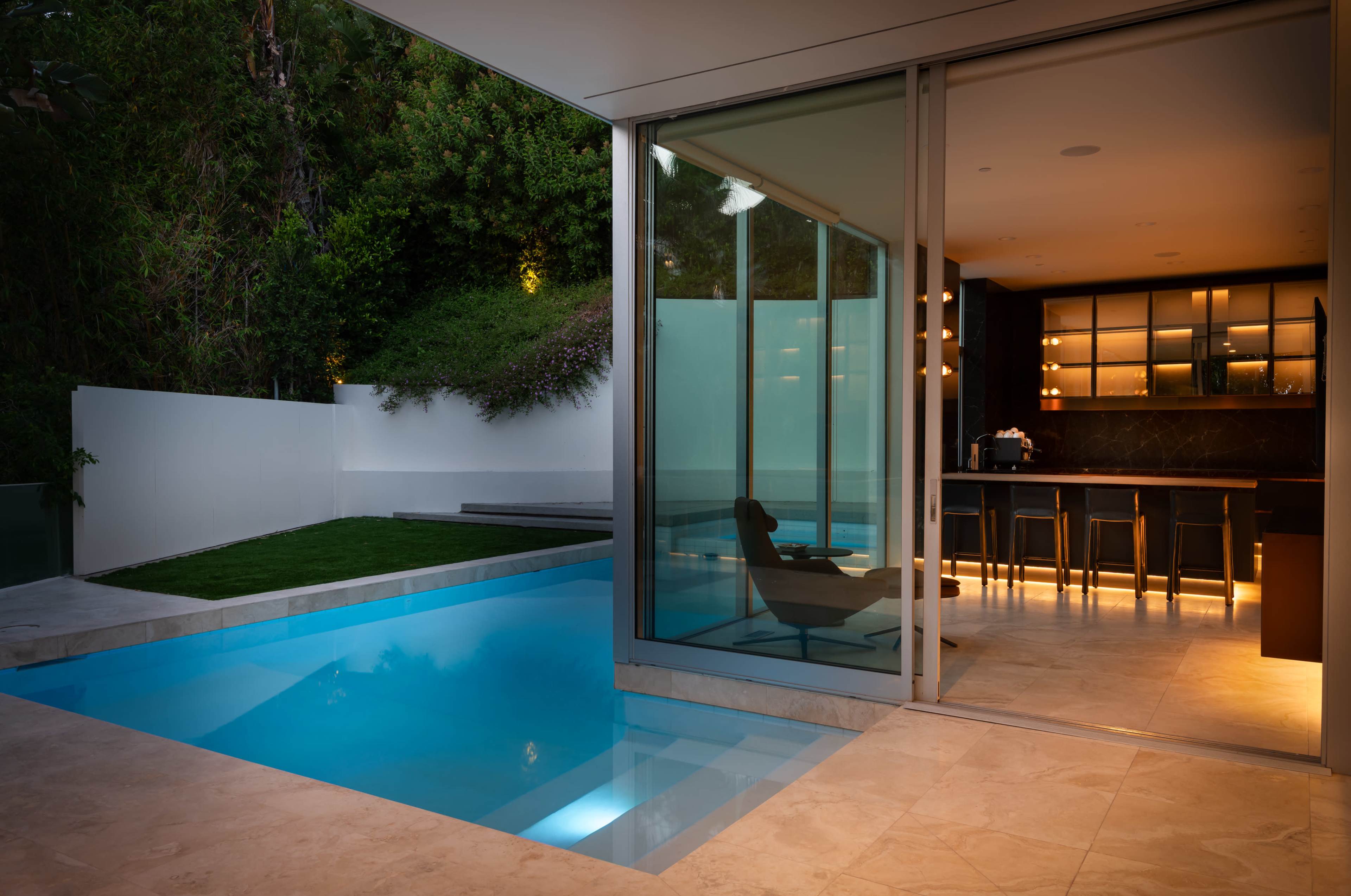 The image shows a modern home with a sleek glass-walled interior that overlooks a swimming pool, surrounded by greenery.