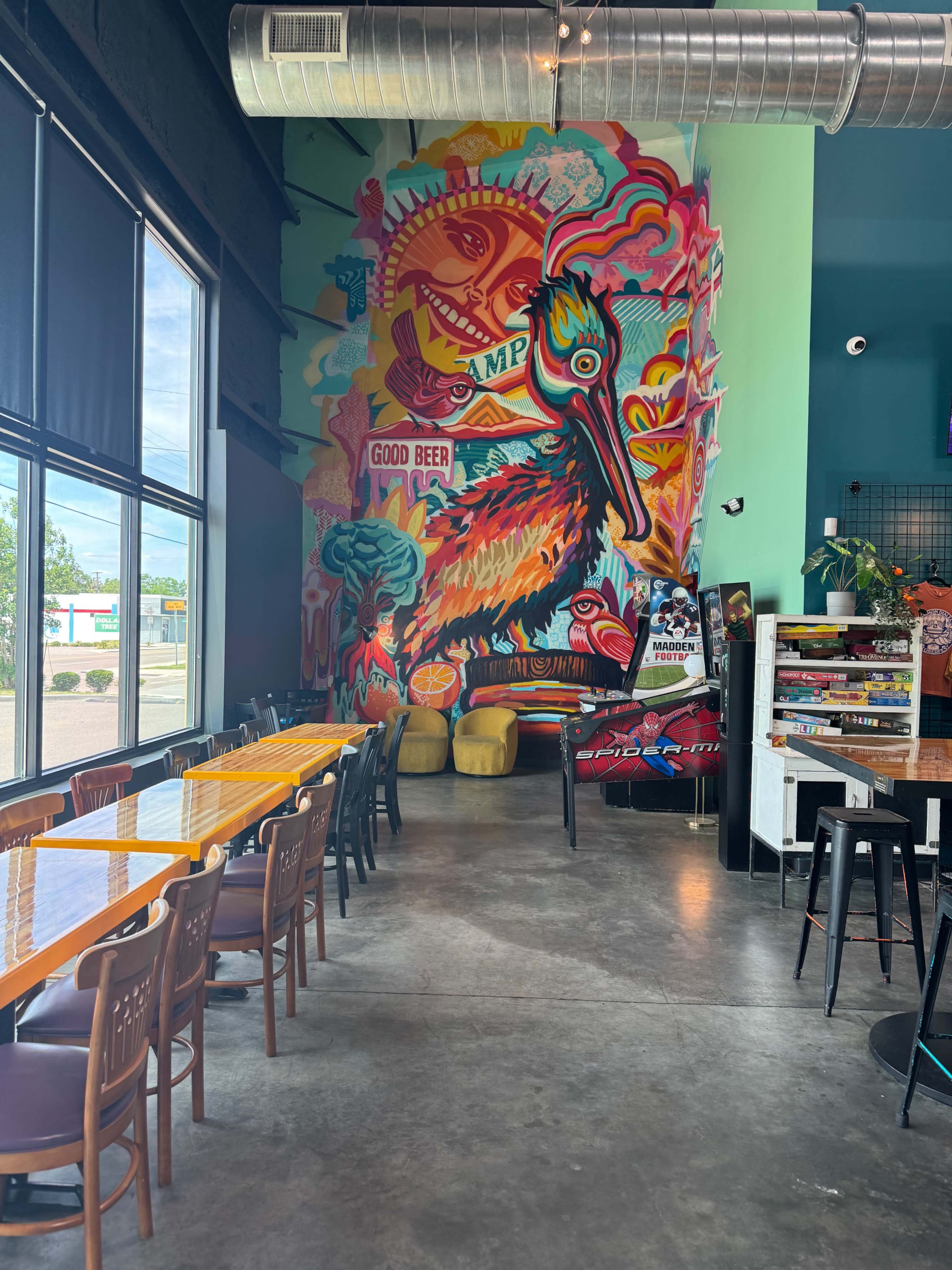 The vibrant interior of a restaurant features a colorful mural of a bird on a wall, with tables and chairs arranged in a row along the space.