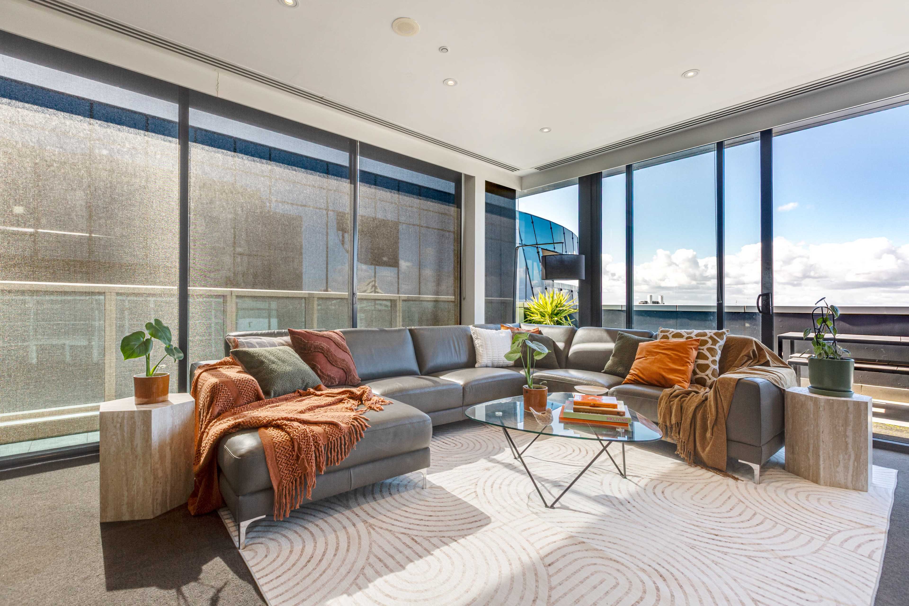 The image shows a modern living room featuring a gray sectional sofa with throw blankets, a glass coffee table, and large windows offering a view of the outdoors.