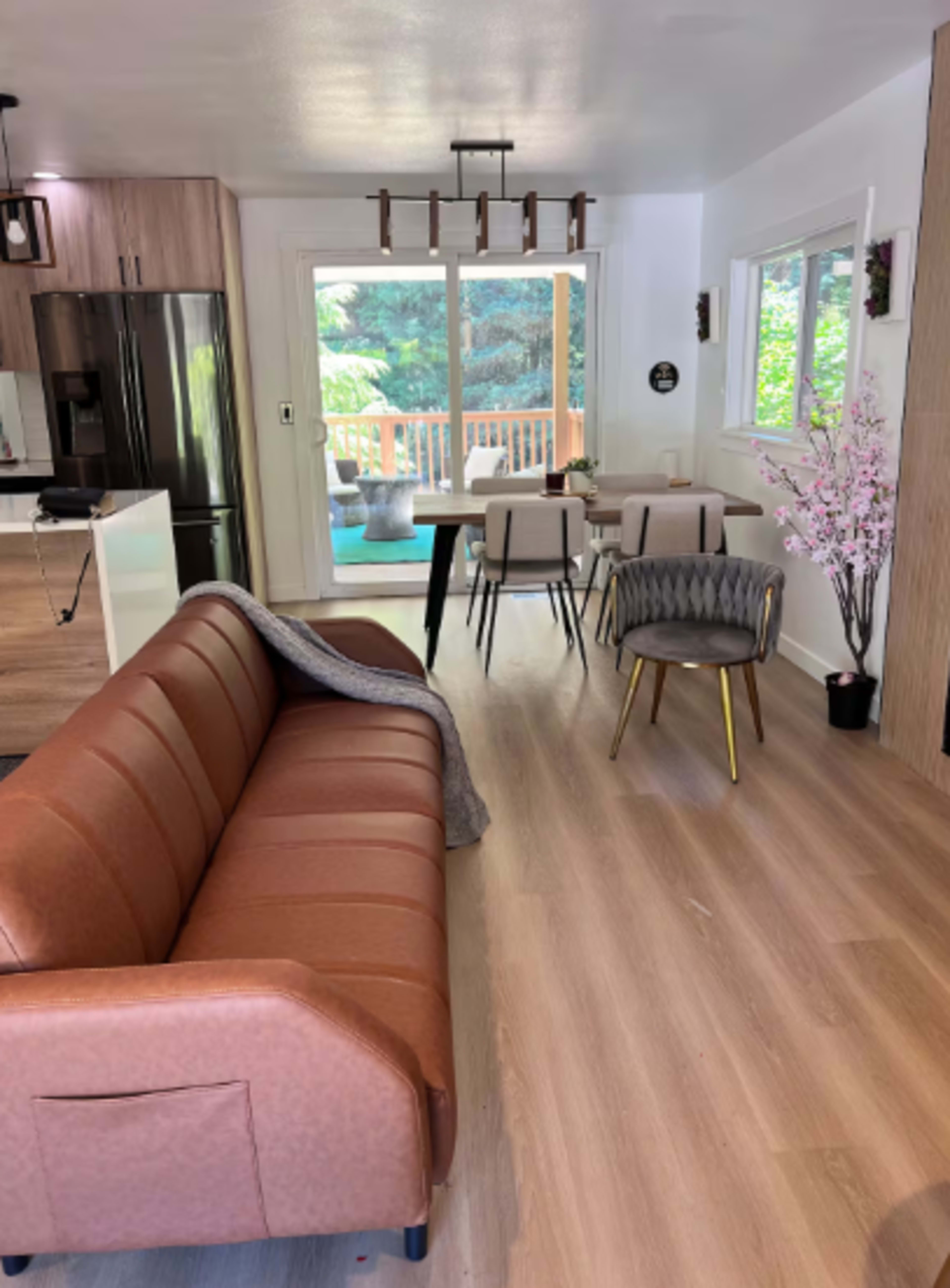 The image shows a modern, open-concept living space featuring a brown sofa, a dining area with a table and chairs, and a large window with a view of a deck and greenery outside.