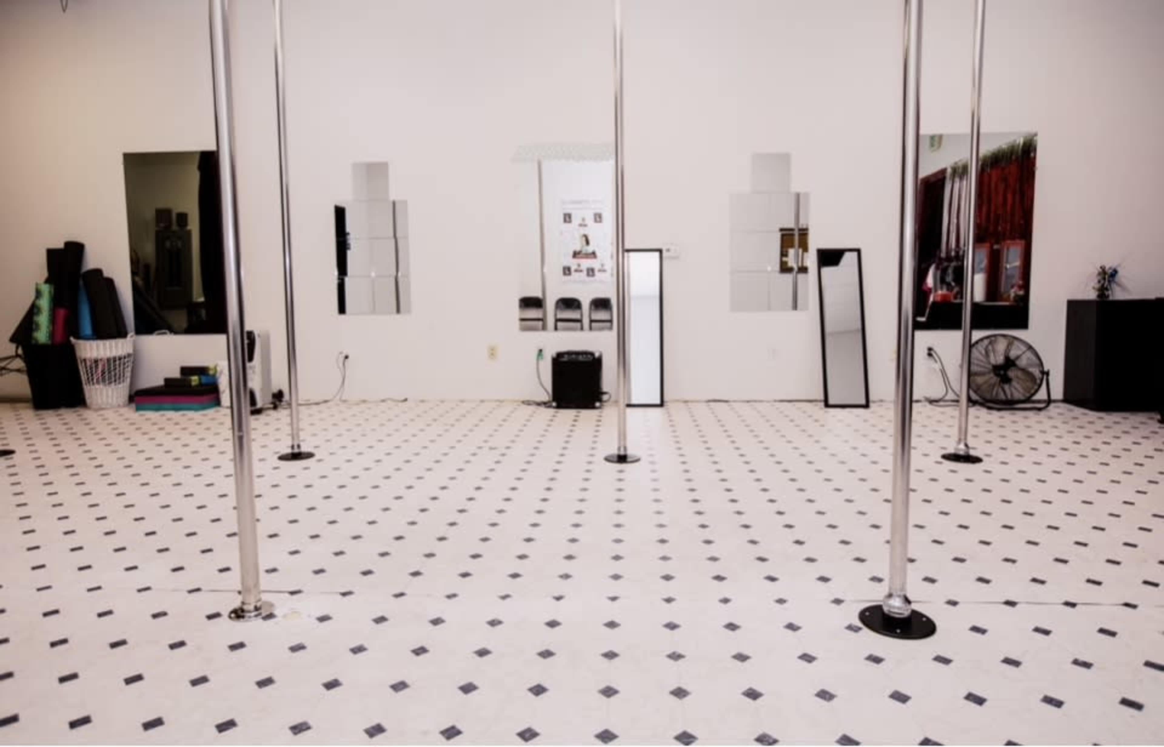 The image shows a spacious dance studio with several vertical poles, a checkered floor, and mirrored walls.