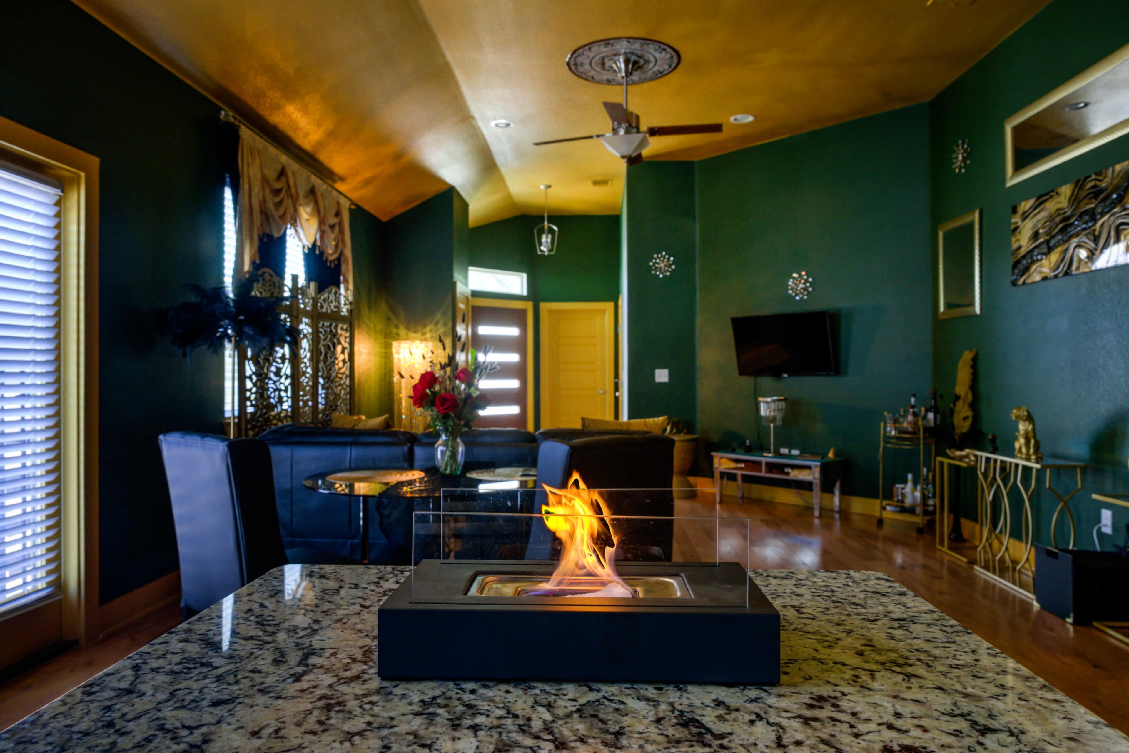 A modern living room features a central decorative fireplace on a granite table, surrounded by dark seating and green walls.