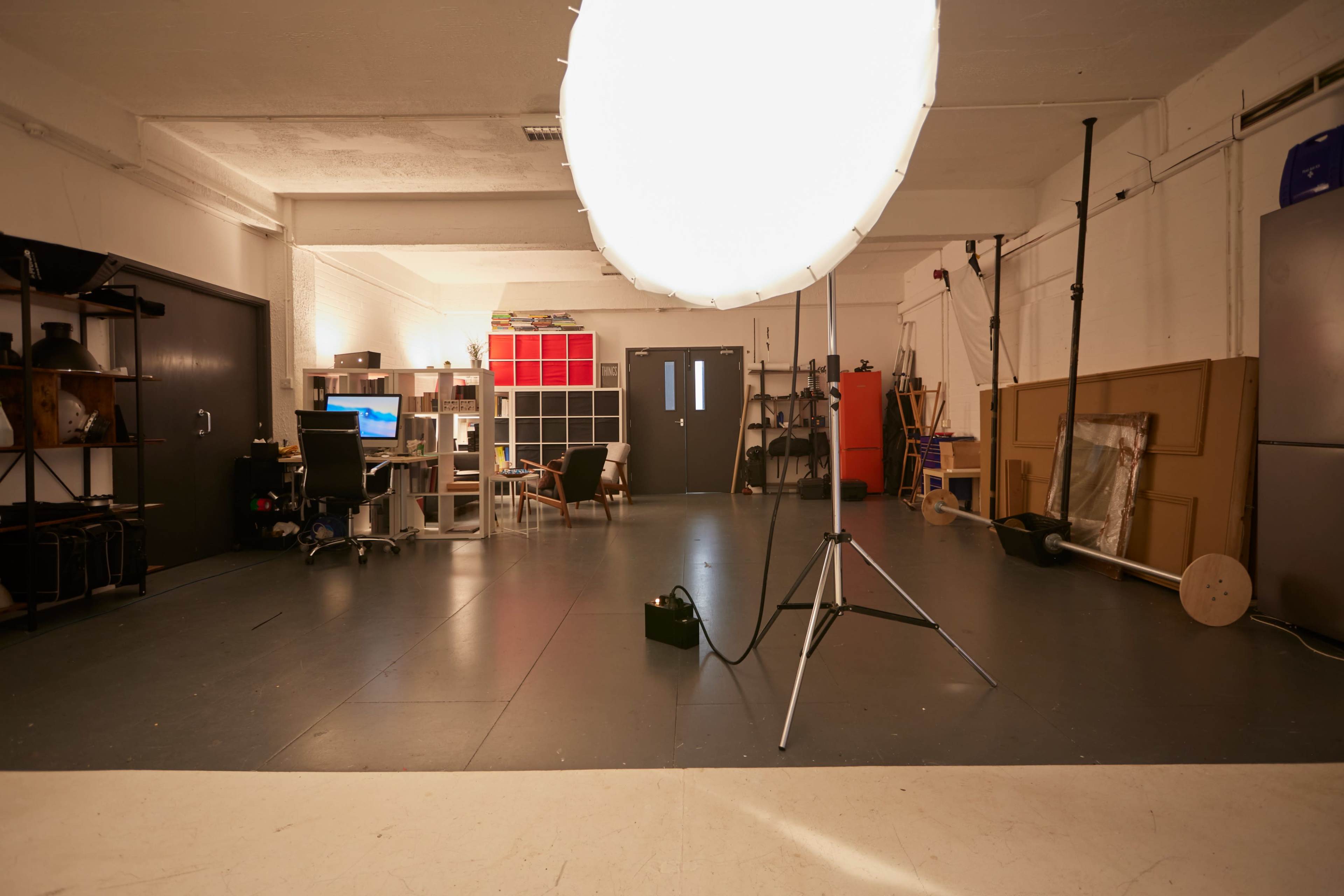 A large lighting setup stands on a tripod in a spacious, well-organized studio with various furniture and equipment.
