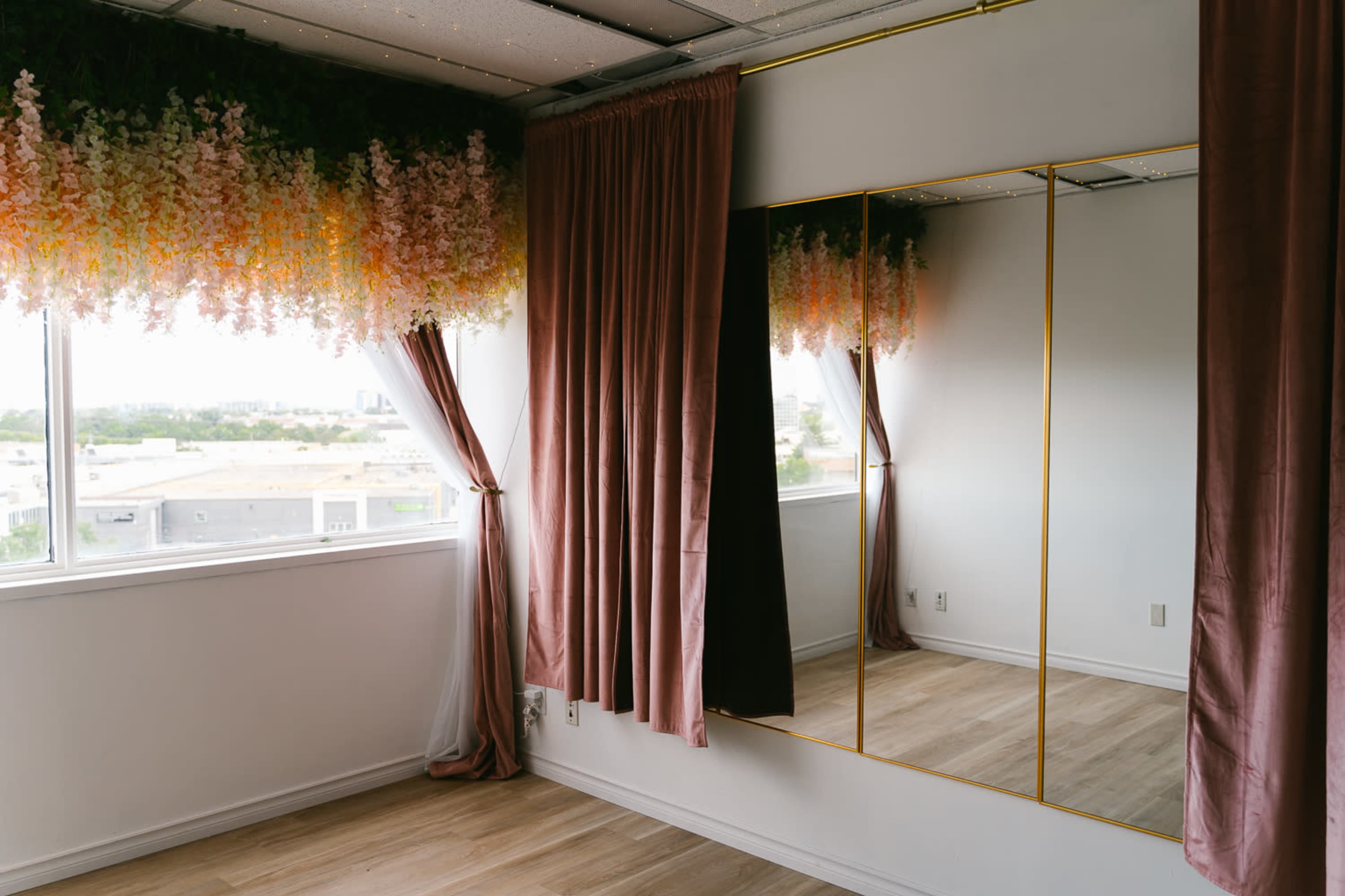 A well-lit room features a large mirror with a gold frame, alongside flowing curtains and a floral arrangement above a window.