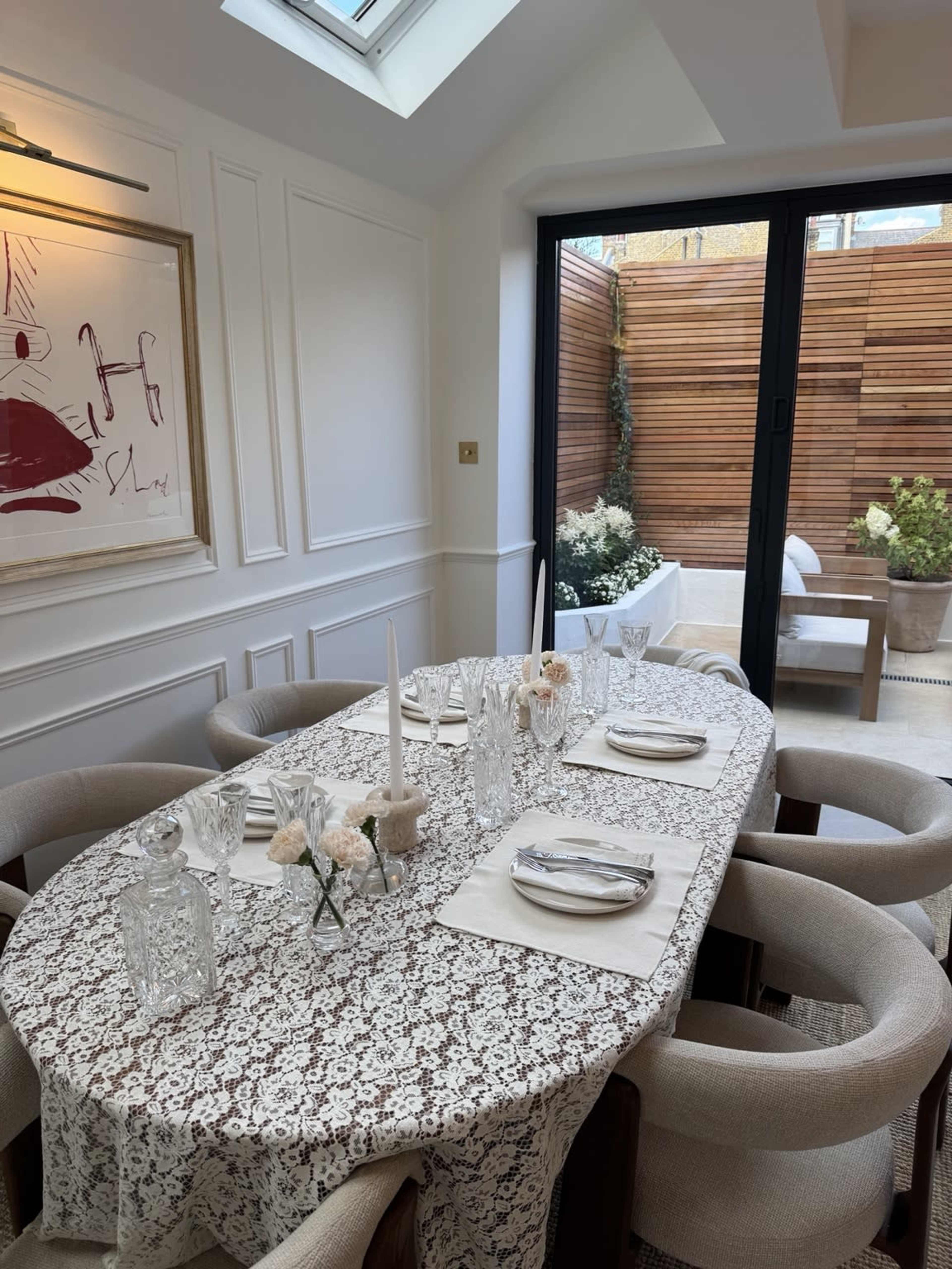 A dining table set with elegant place settings and a tablecloth is arranged in a bright room with a view of a private outdoor space.