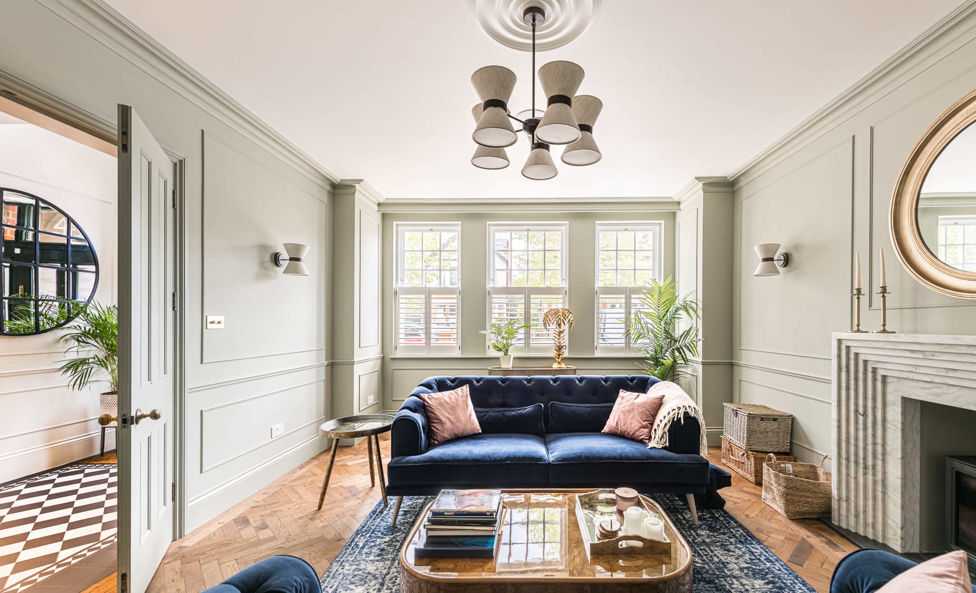 The image shows a stylish living room with a blue velvet sofa, large windows, a decorative mirror, and a wooden floor.