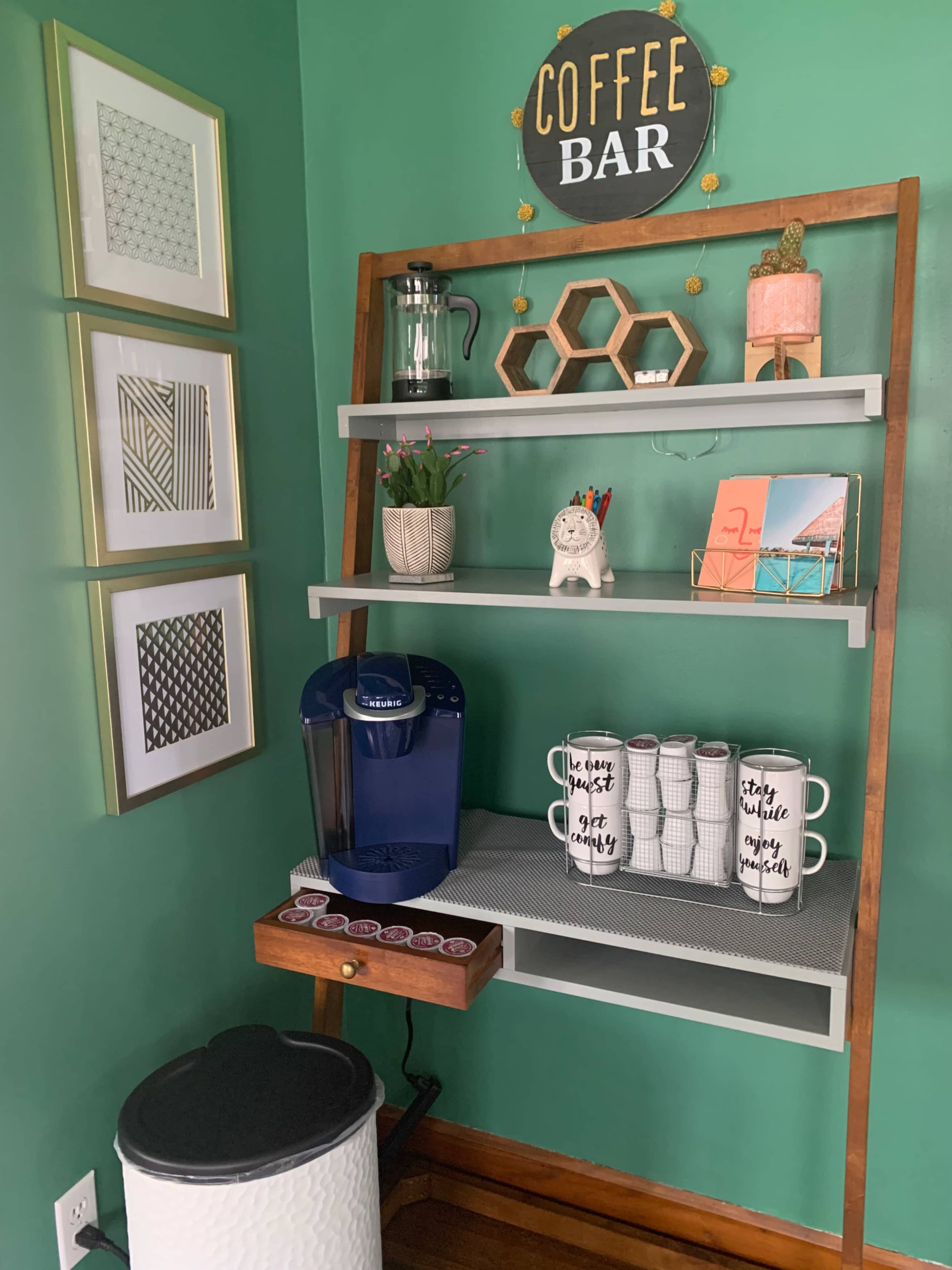 A wooden coffee bar setup features shelves with decorative items, a coffee maker, mugs, and a small trash can against a green wall.
