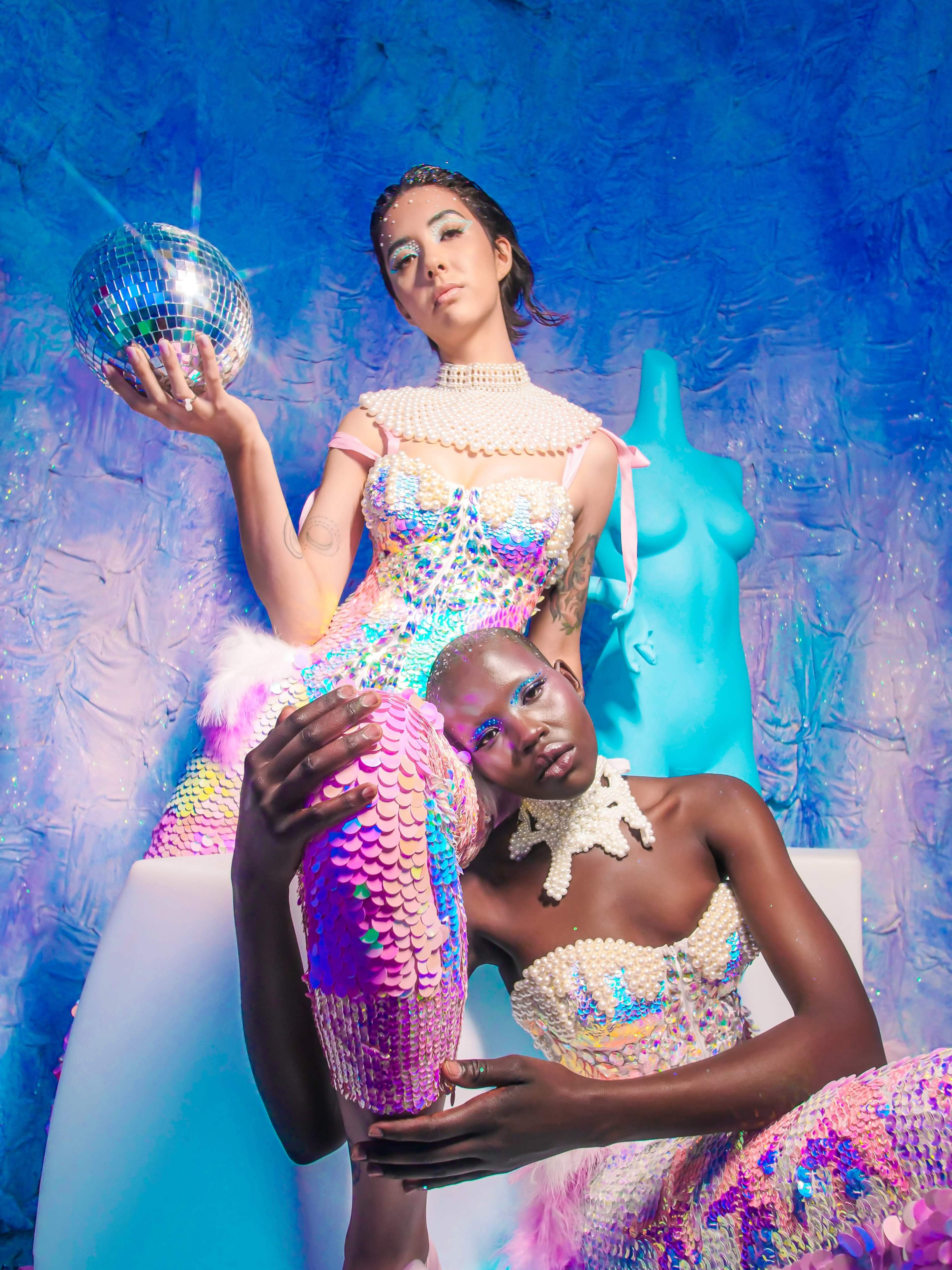 Two models pose with a disco ball and a mannequin against a vibrant, textured blue background, showcasing shimmering, sequined outfits.