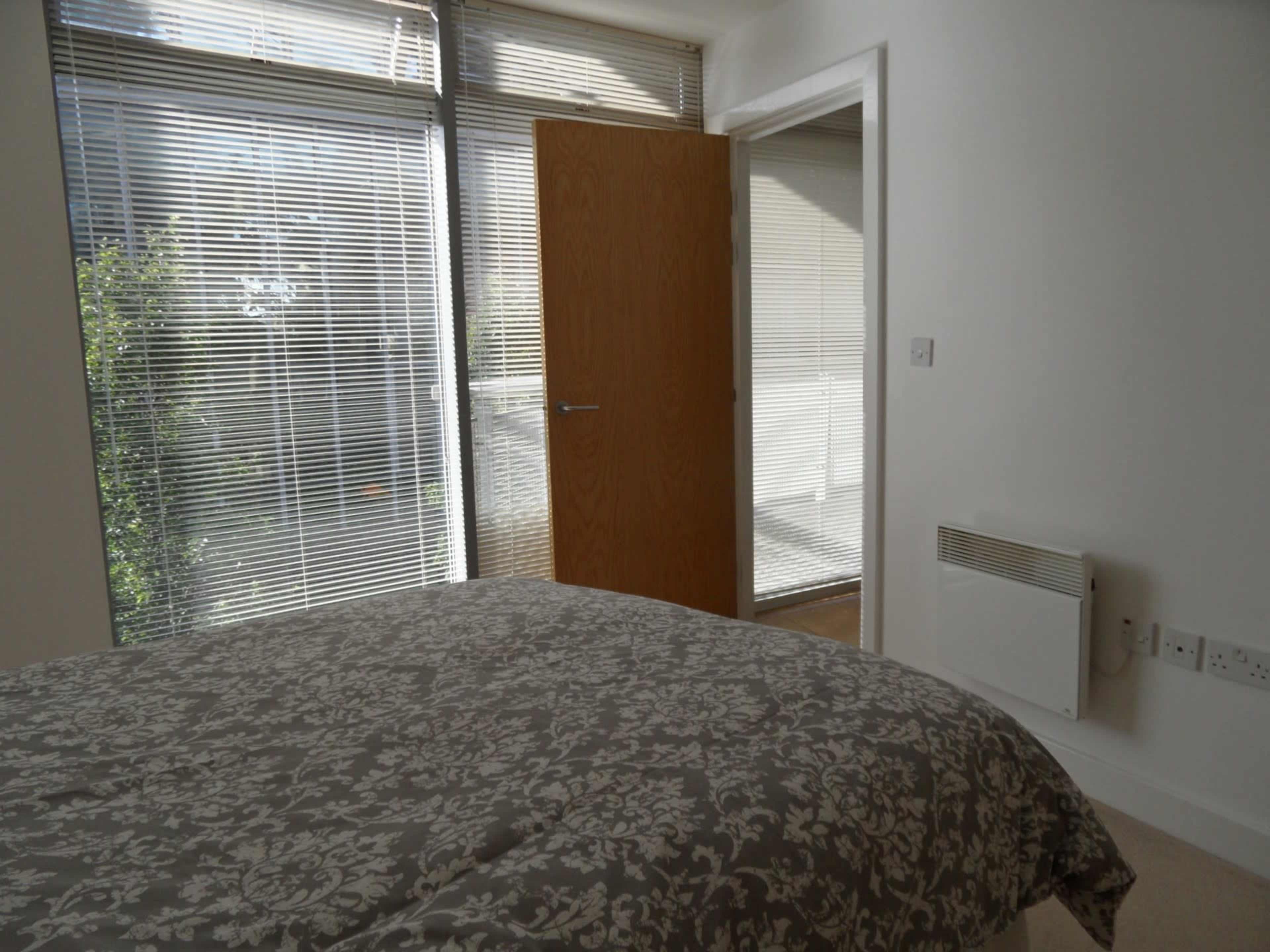 A bedroom features a bed with a patterned duvet, a door leading to a balcony, and large windows covered with blinds.