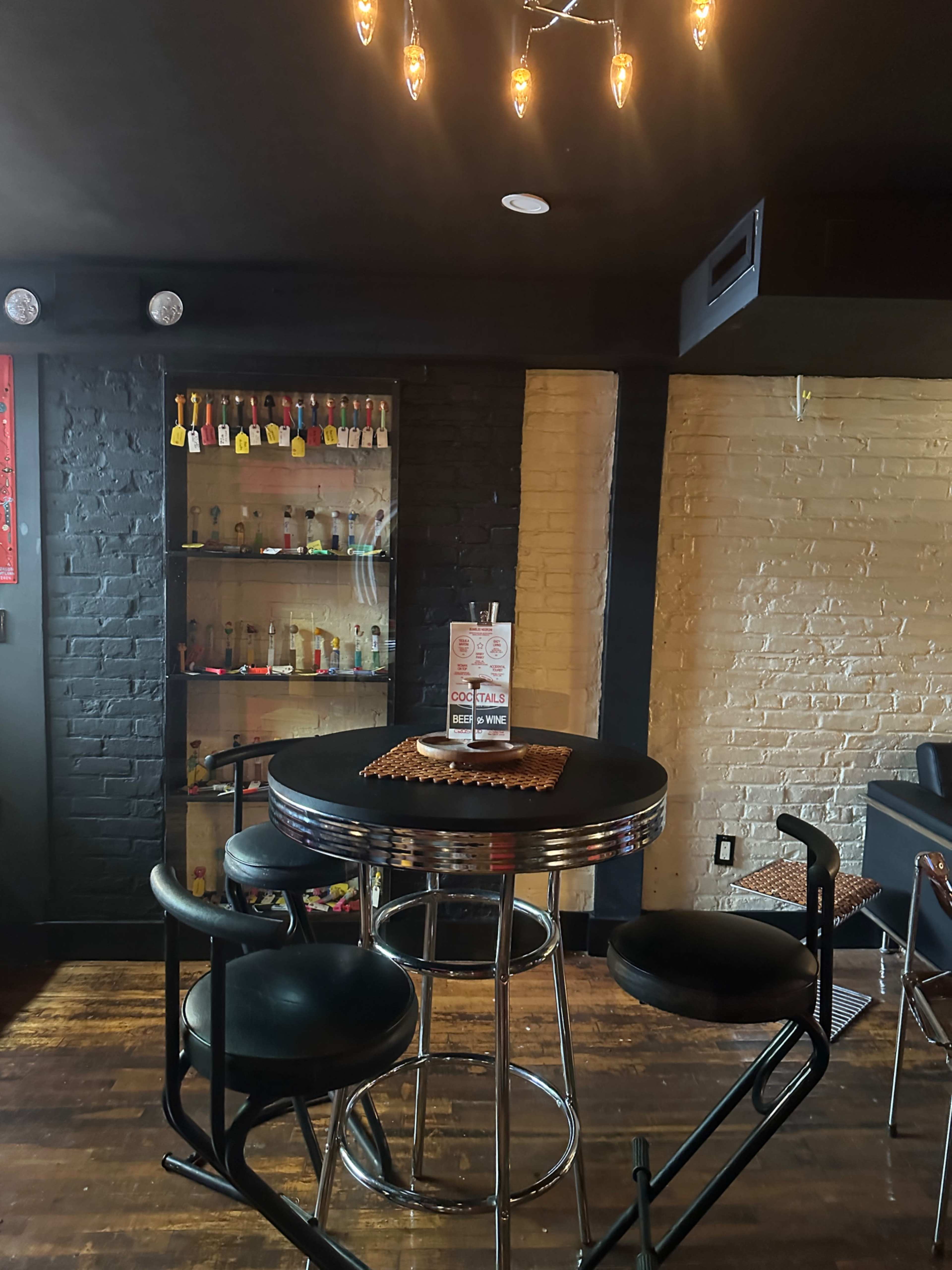 A round bar table with black stools is positioned against a backdrop of black and yellow-brick walls, featuring a display shelf stocked with various bottles and cocktail menus on the table.