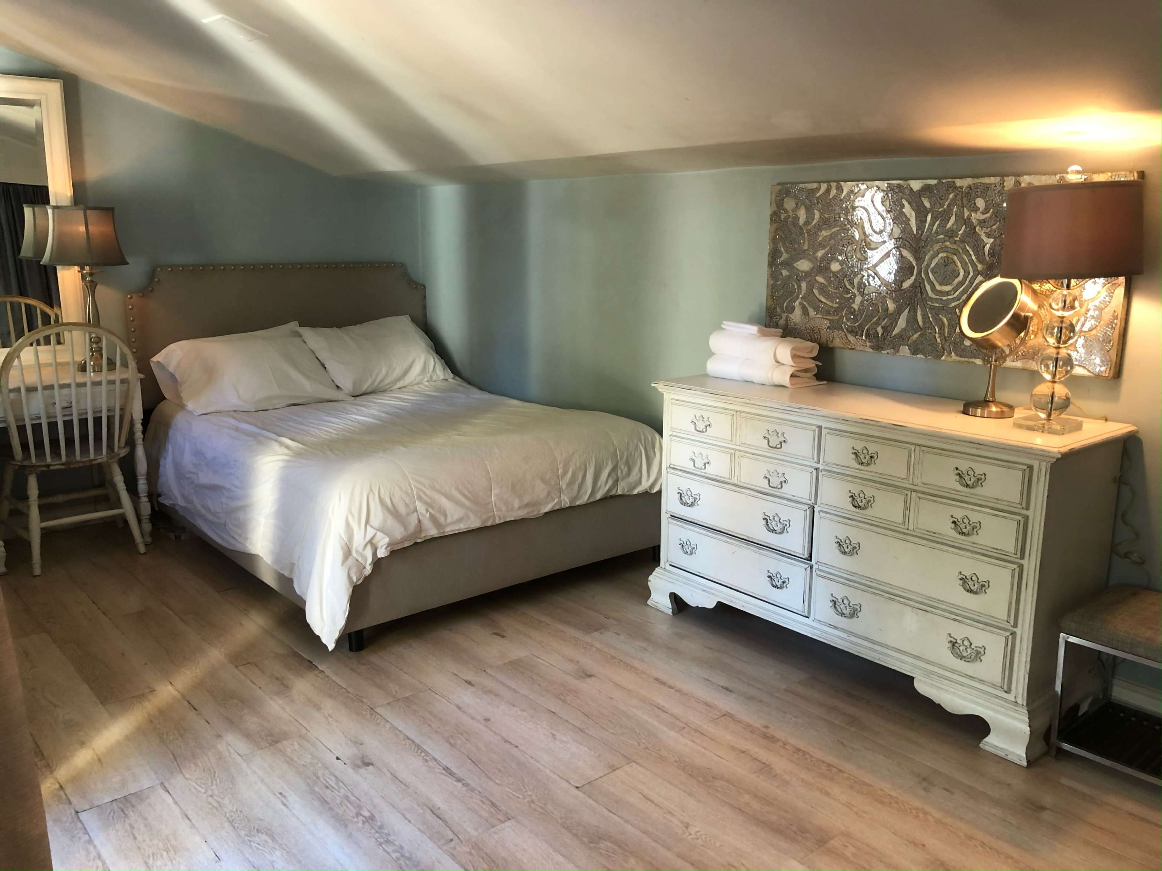 The image shows a cozy attic room with a bed, a dresser, a chair, and a lamp, all set against light blue walls.