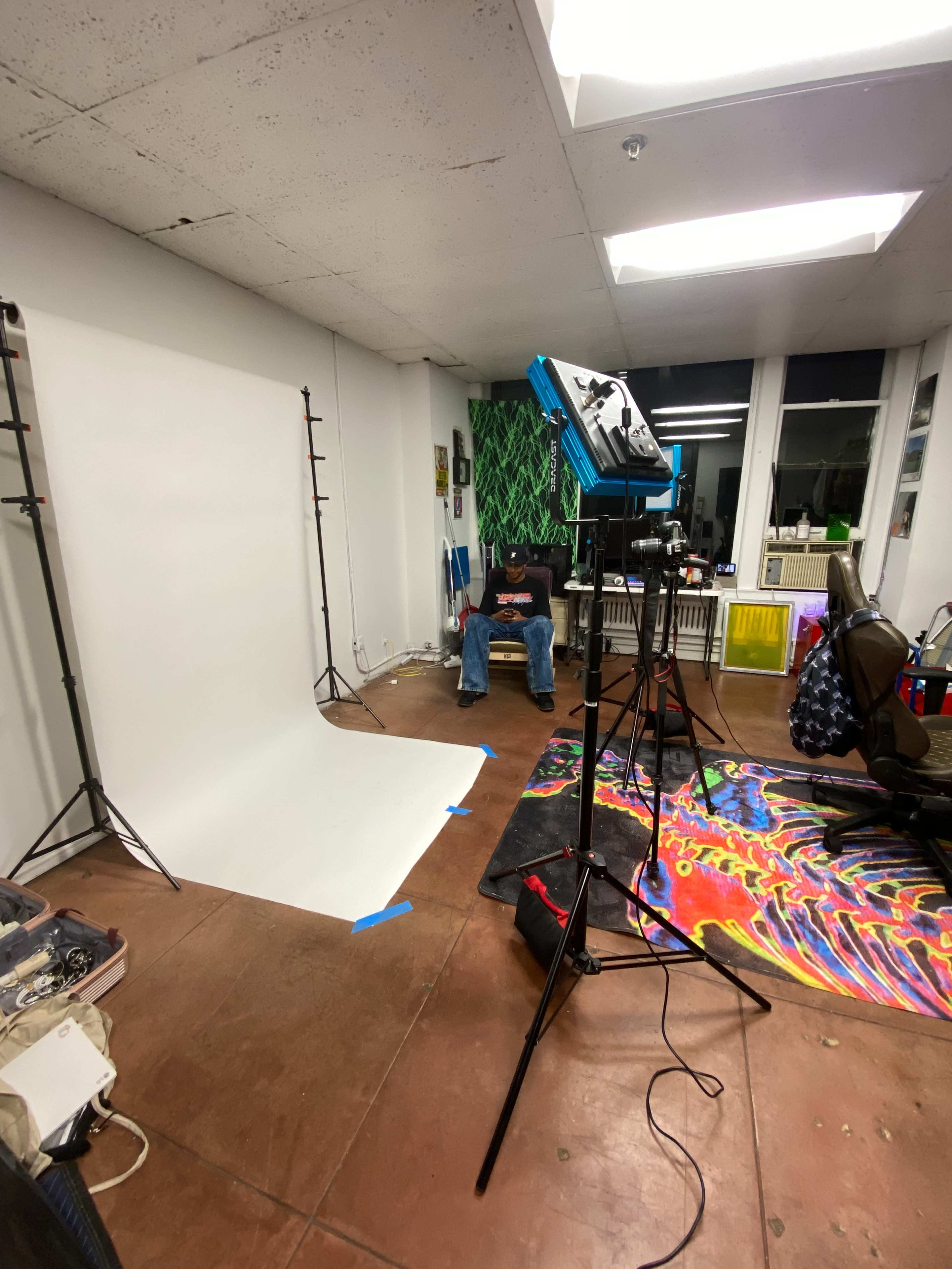 A man sits comfortably in a chair in a photo studio with a white backdrop, studio lights, and colorful carpets scattered on the floor.