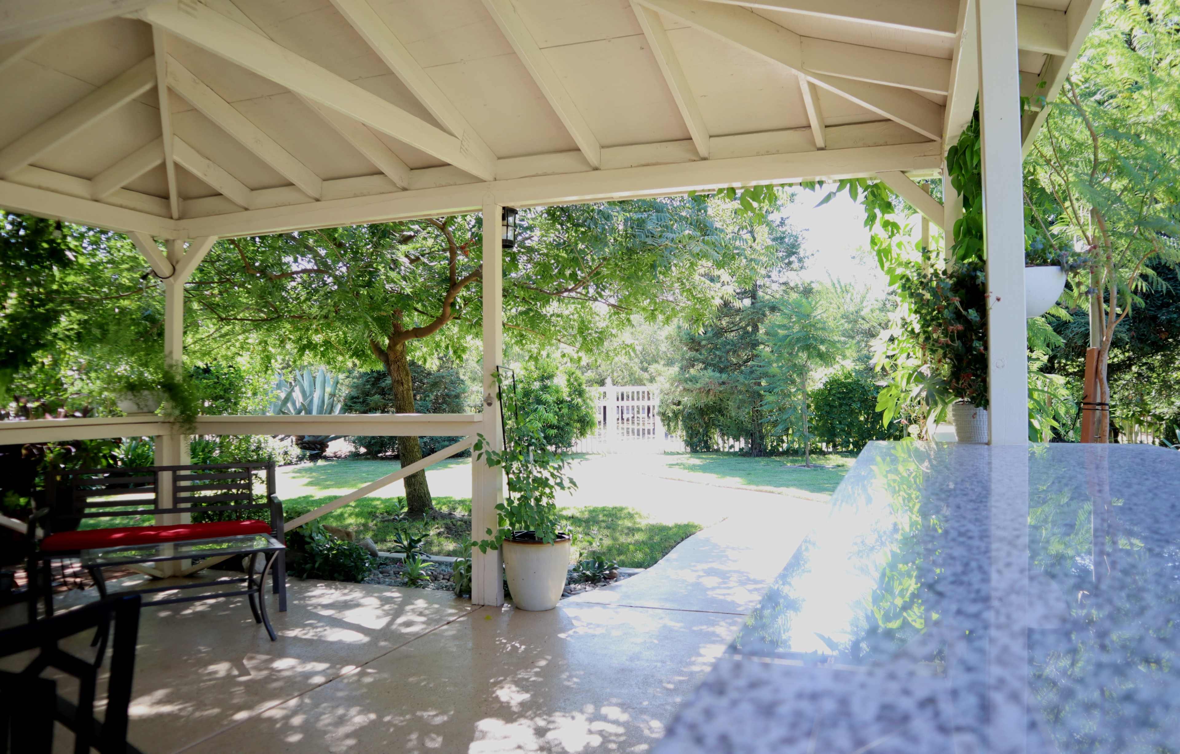 A covered patio features a seating area and overlooks a landscaped garden with trees and a path.
