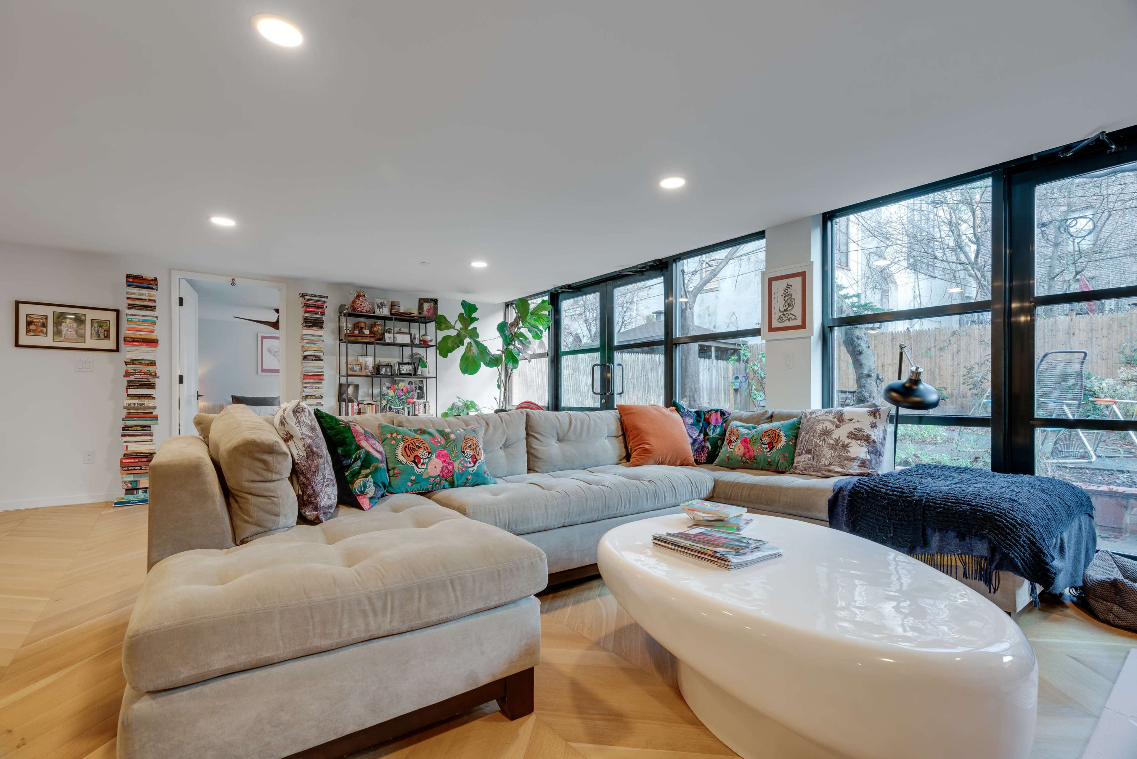 A modern living room with a large beige sectional sofa, colorful decorative pillows, a white coffee table, and large windows allowing natural light.
